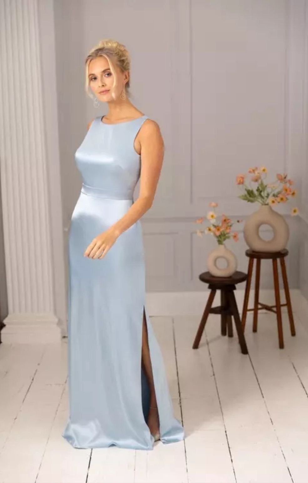 Woman in a long, light blue satin evening gown with a slit, standing indoors with flowers in vases on small wooden tables behind her.