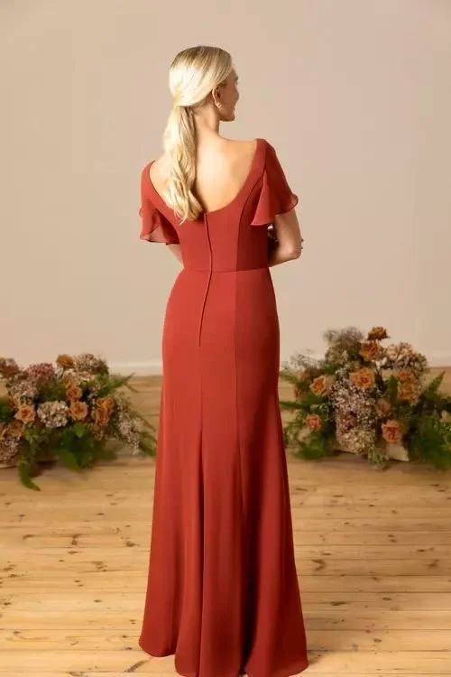A woman with blonde hair wearing a long, red gown with flutter sleeves, standing with her back to the camera. There are flowers and greenery on the floor behind her.