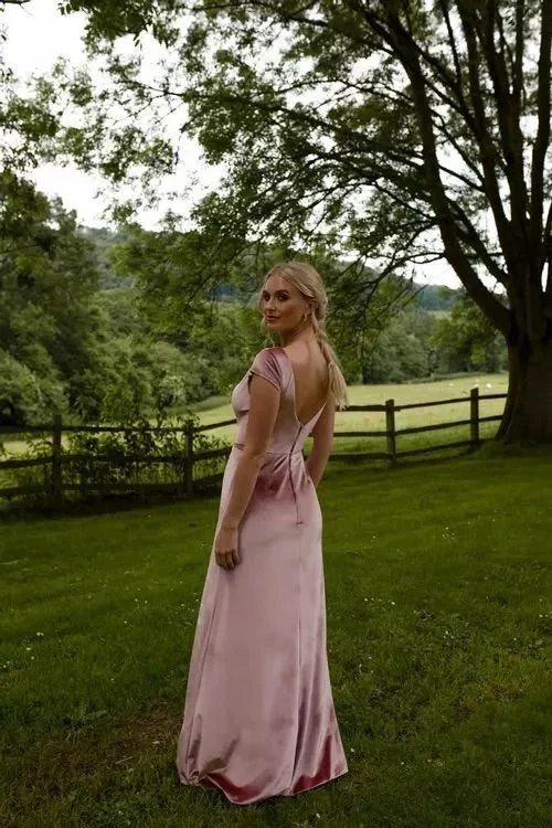 A woman wearing a pink satin gown standing in a grassy field with trees and a wooden fence in the background.