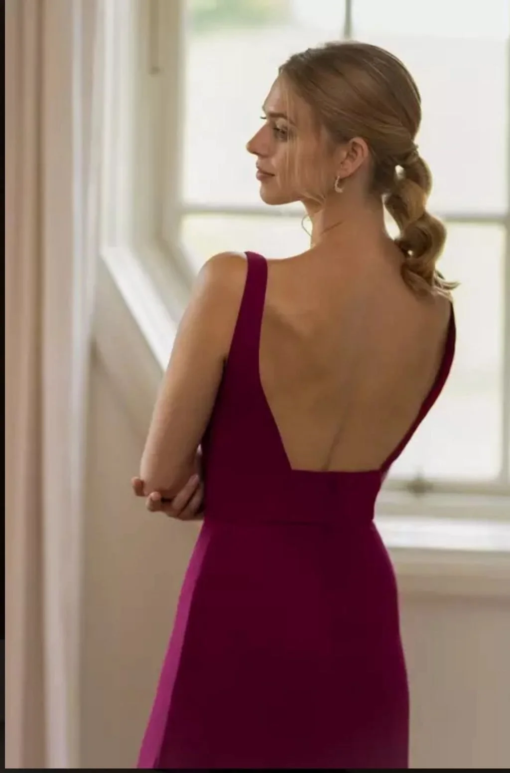 A woman with reddish-brown hair styled in loose curls, wearing a sleeveless, deep red gown with an open back, standing indoors near a window with natural light.