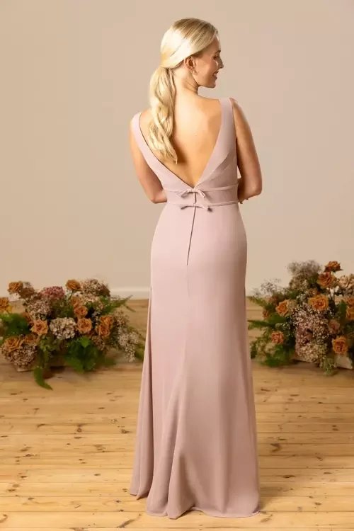 A woman with long blonde hair wearing a light pink, sleeveless, floor-length gown with a deep V back, standing on a wooden floor surrounded by flower arrangements.