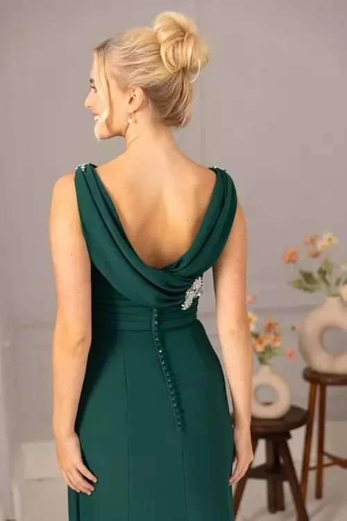 Woman in a dark green elegant dress with a deep back and a decorative brooch, smiling and standing in a room with floral decor.