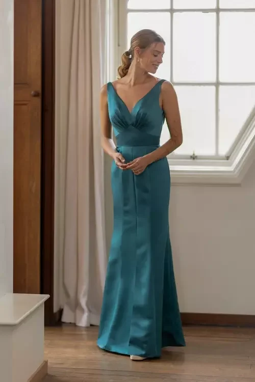 A woman in a teal evening gown standing in front of a window, with beige curtains and wooden flooring.