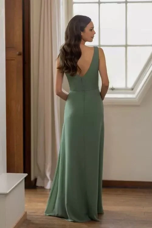 Woman in a long green dress standing indoors near a window, with her back to the camera and hands behind her back.