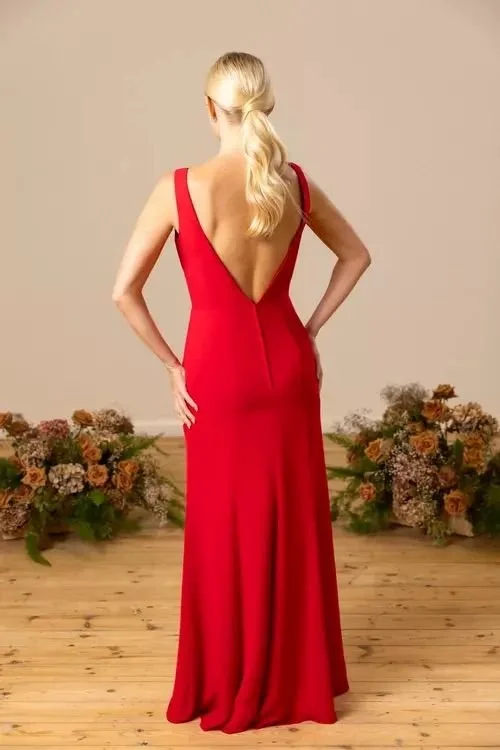 Back view of a woman wearing a long, elegant red dress with a deep V back, standing on a wooden floor with flower arrangements in the background.