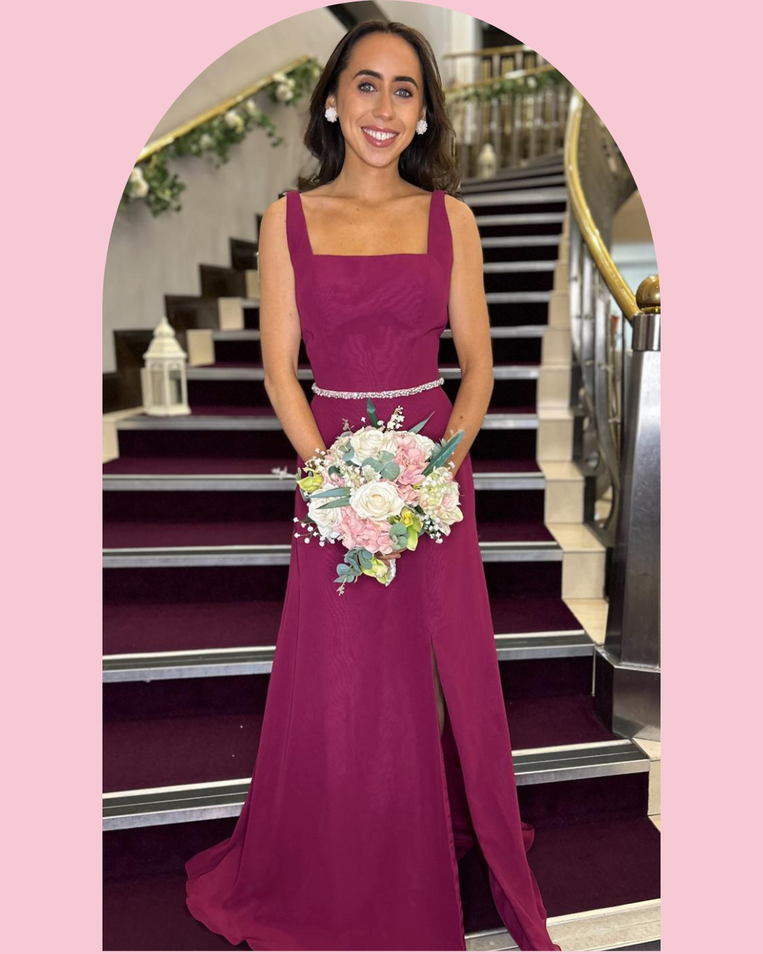 A woman in a magenta gown holding a bouquet of roses and greenery, standing on a staircase inside a decorated venue.
