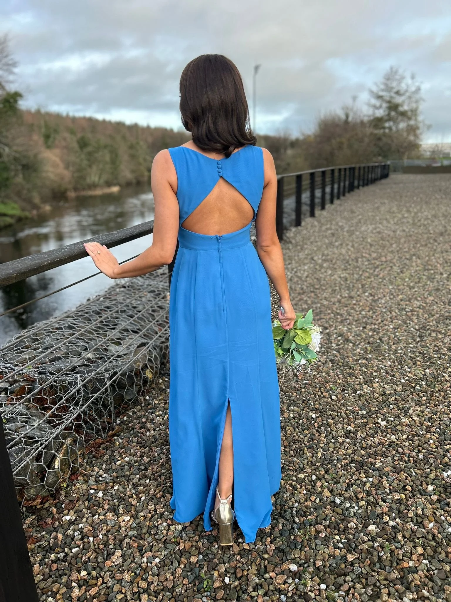 Woman in a long blue dress holding a bouquet, standing on a gravel path near a railing beside a river, with trees and cloudy sky in the background.