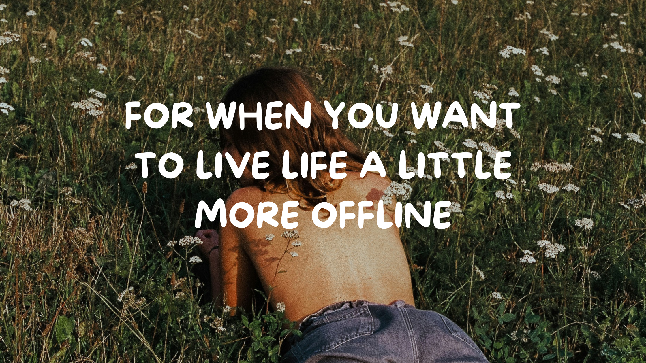 A person with brown hair and tattoos lying face down in a grassy field with small white flowers, wearing denim shorts.