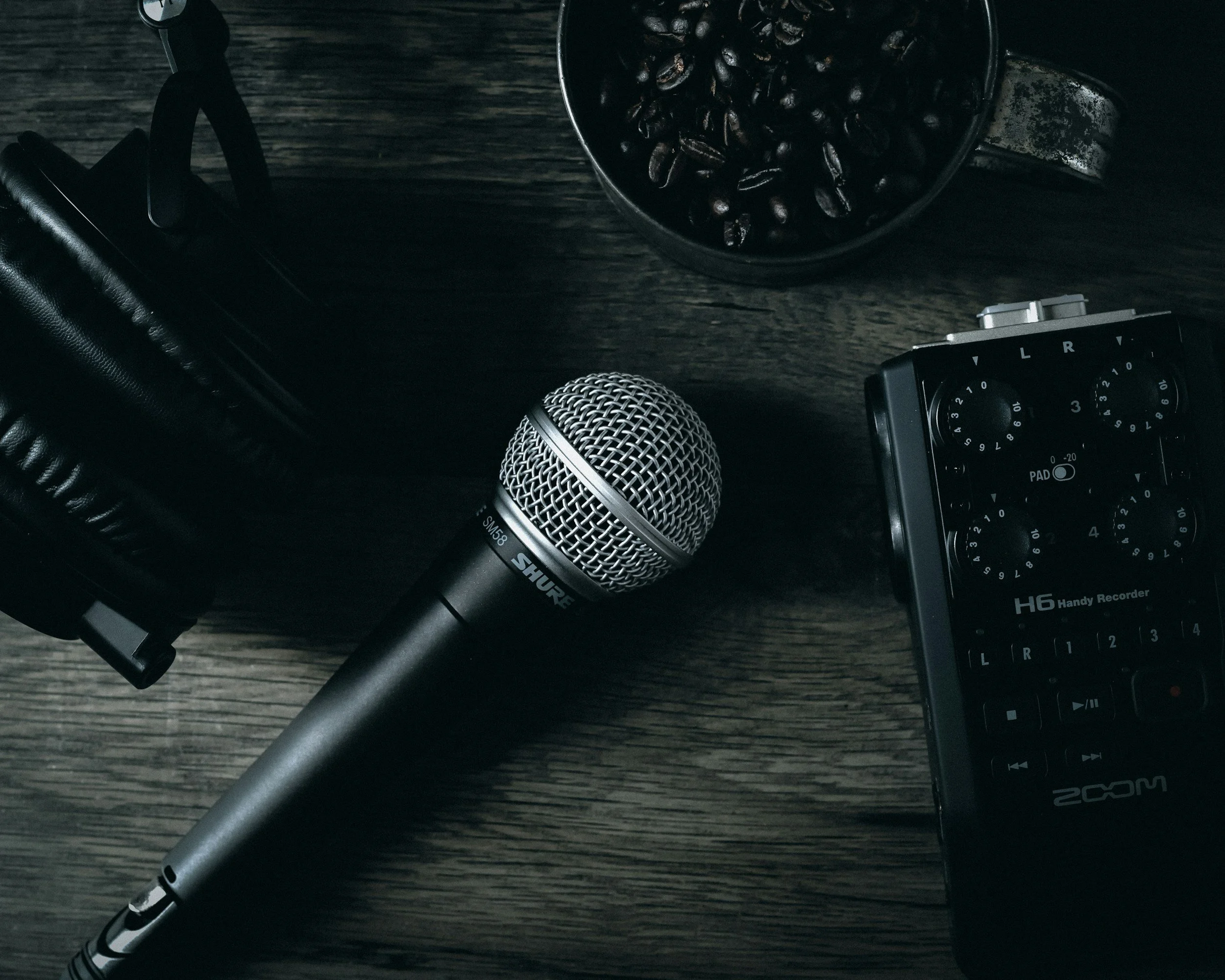 A microphone, a pair of headphones, a cup with coffee beans, and a portable digital recorder are arranged on a wooden surface.