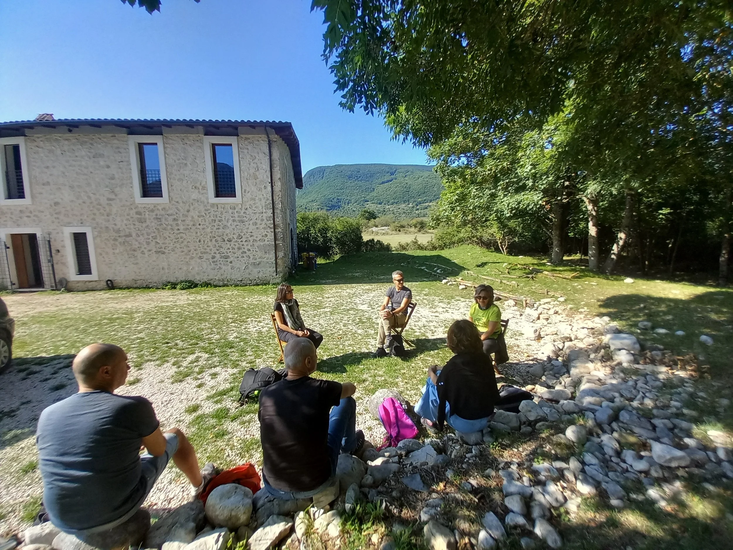 Group of people sitting outdoors in a circle under trees on a sunny day, with a stone building and mountains in the background.