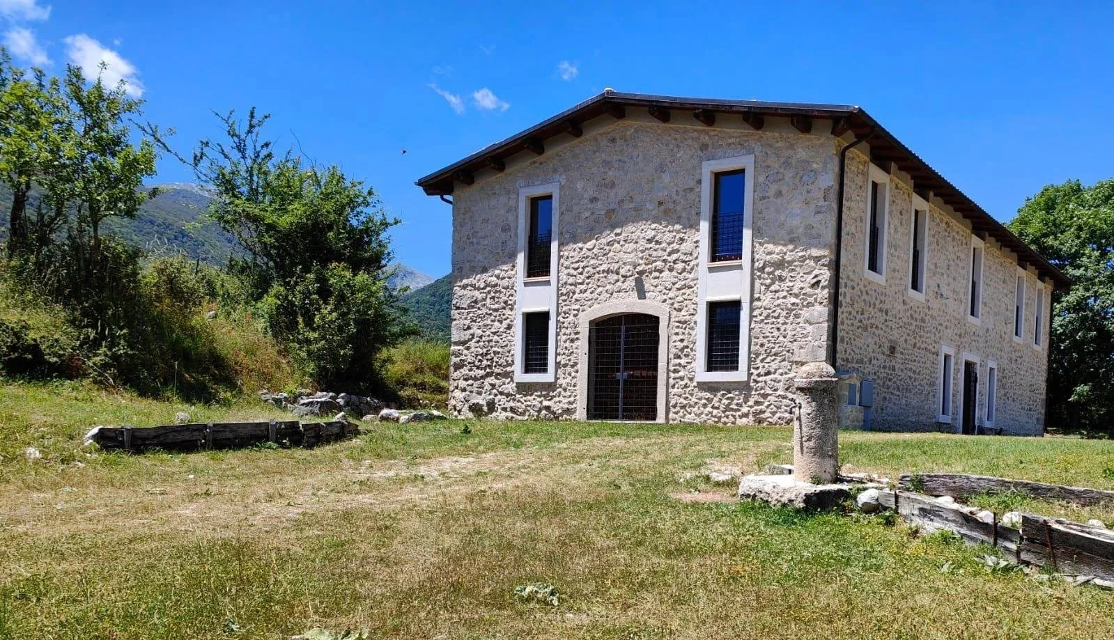 A two-story stone house with multiple narrow windows, set on a grassy landscape with trees and mountains in the background, on a clear sunny day.