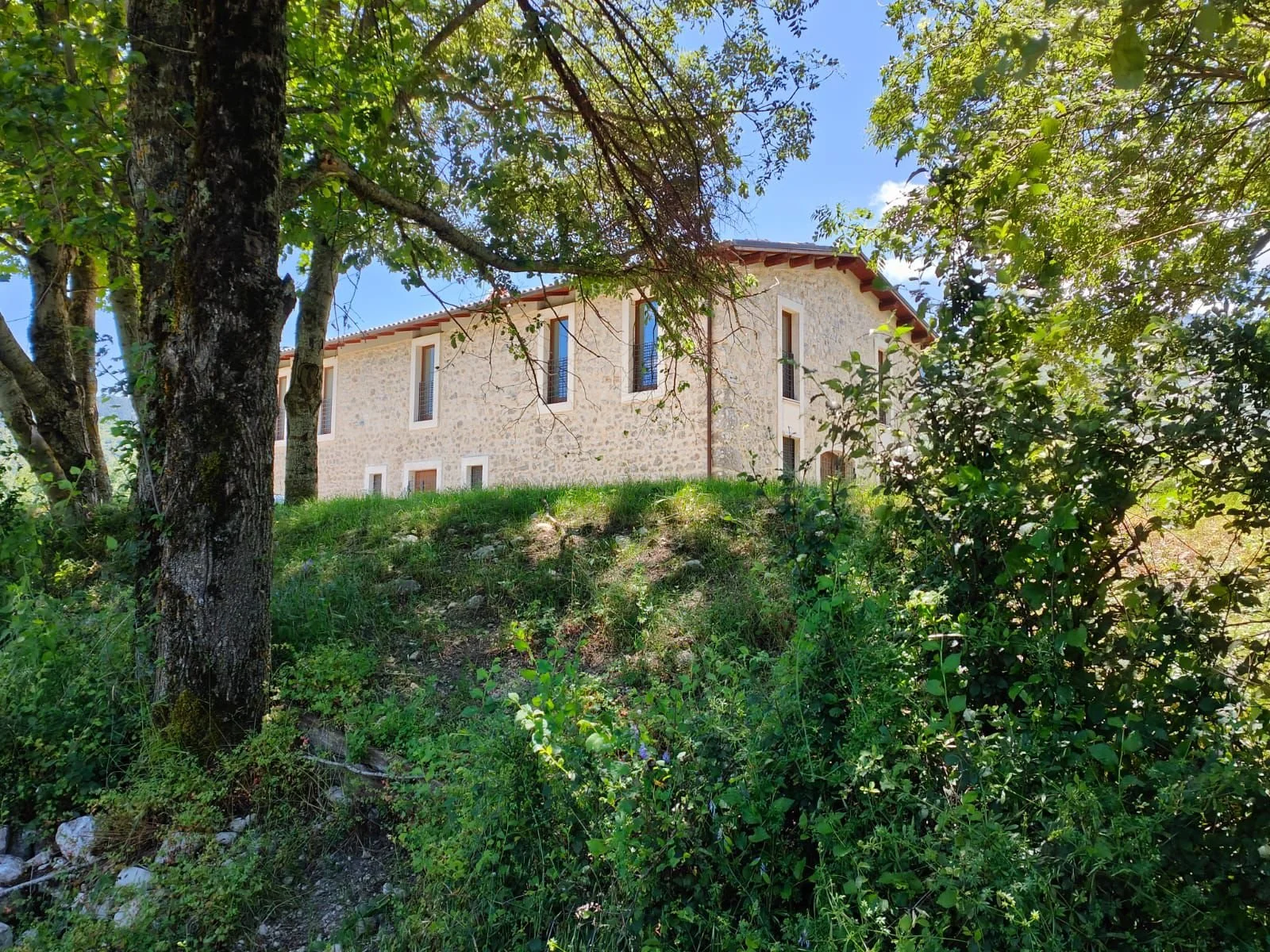 Stone house on a grassy hill surrounded by trees and bushes under a blue sky.