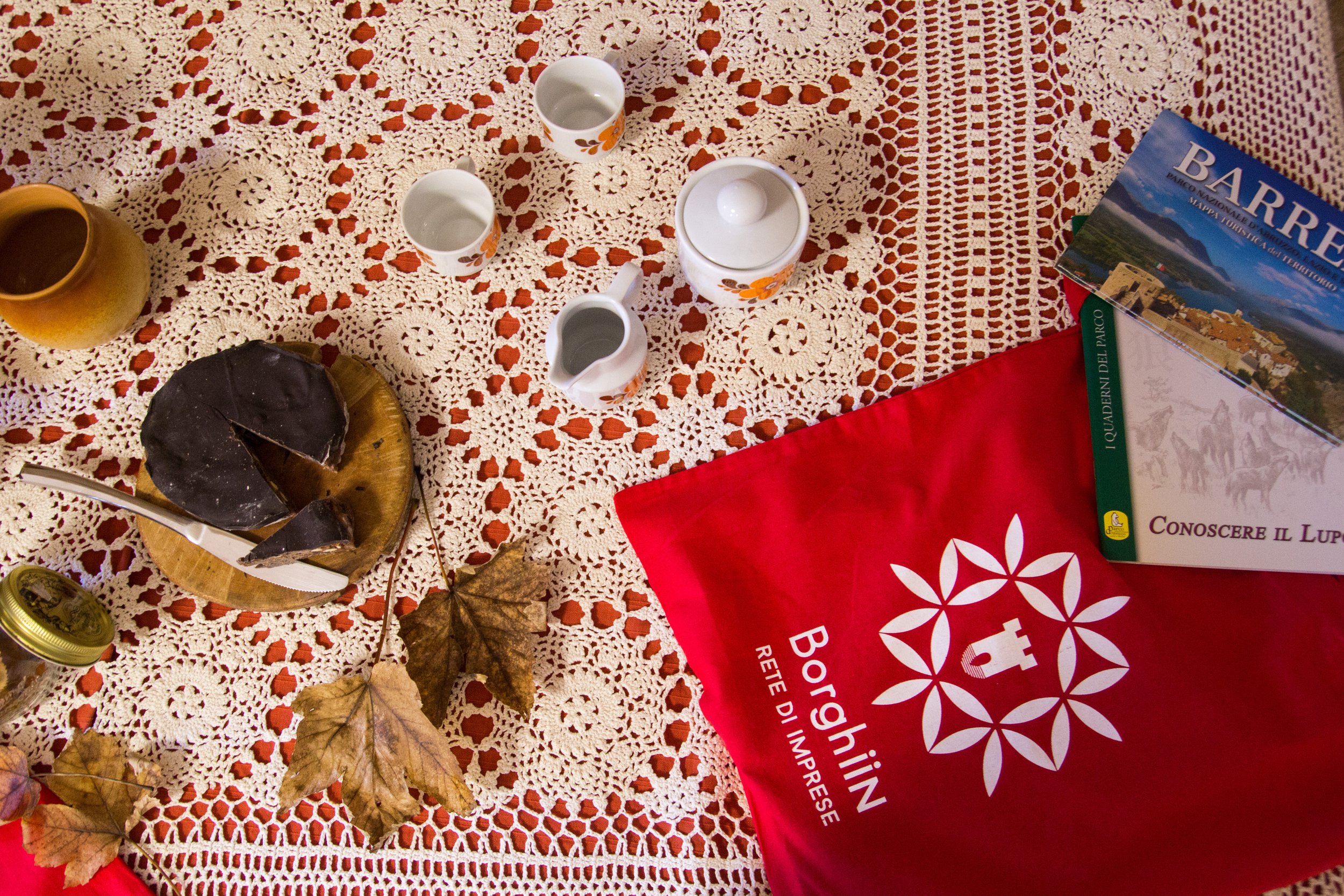 Table with a red cloth displaying the logo of Bighin, three books including one about Barrea, cups, a teapot, and a chocolate dessert with leaves placed on a lace tablecloth.