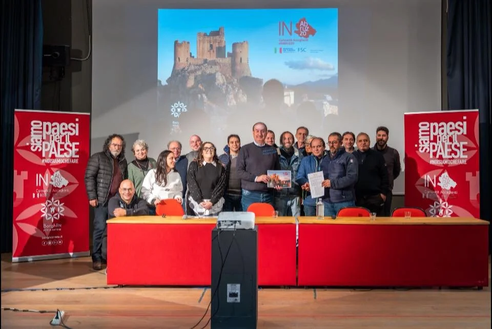 Group of people standing and sitting behind a table on a stage, with a large screen displaying a castle and text in the background, and banners on either side of the stage.
