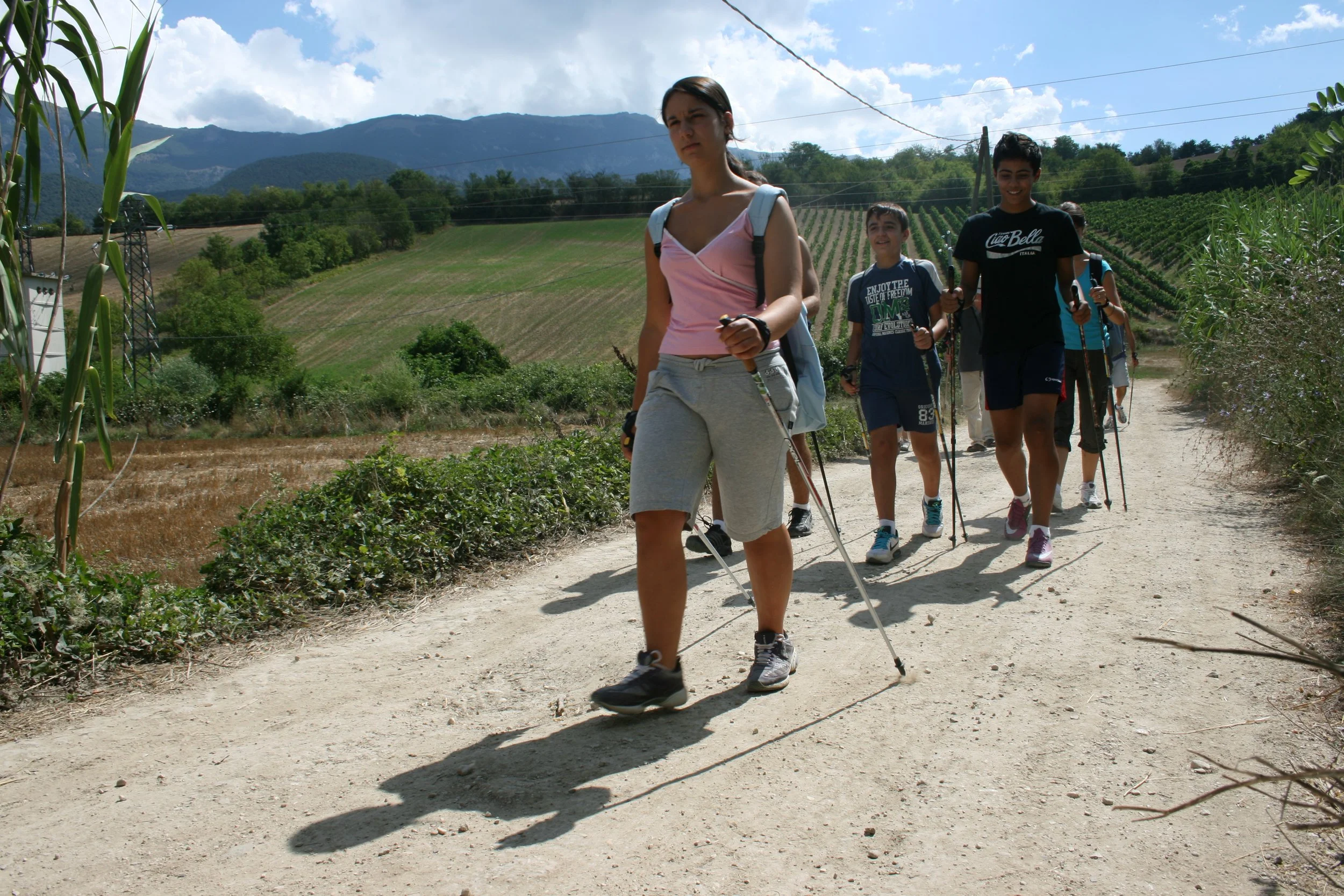 Group of young people hiking on a dirt trail in a rural area with hills and farmland in the background.