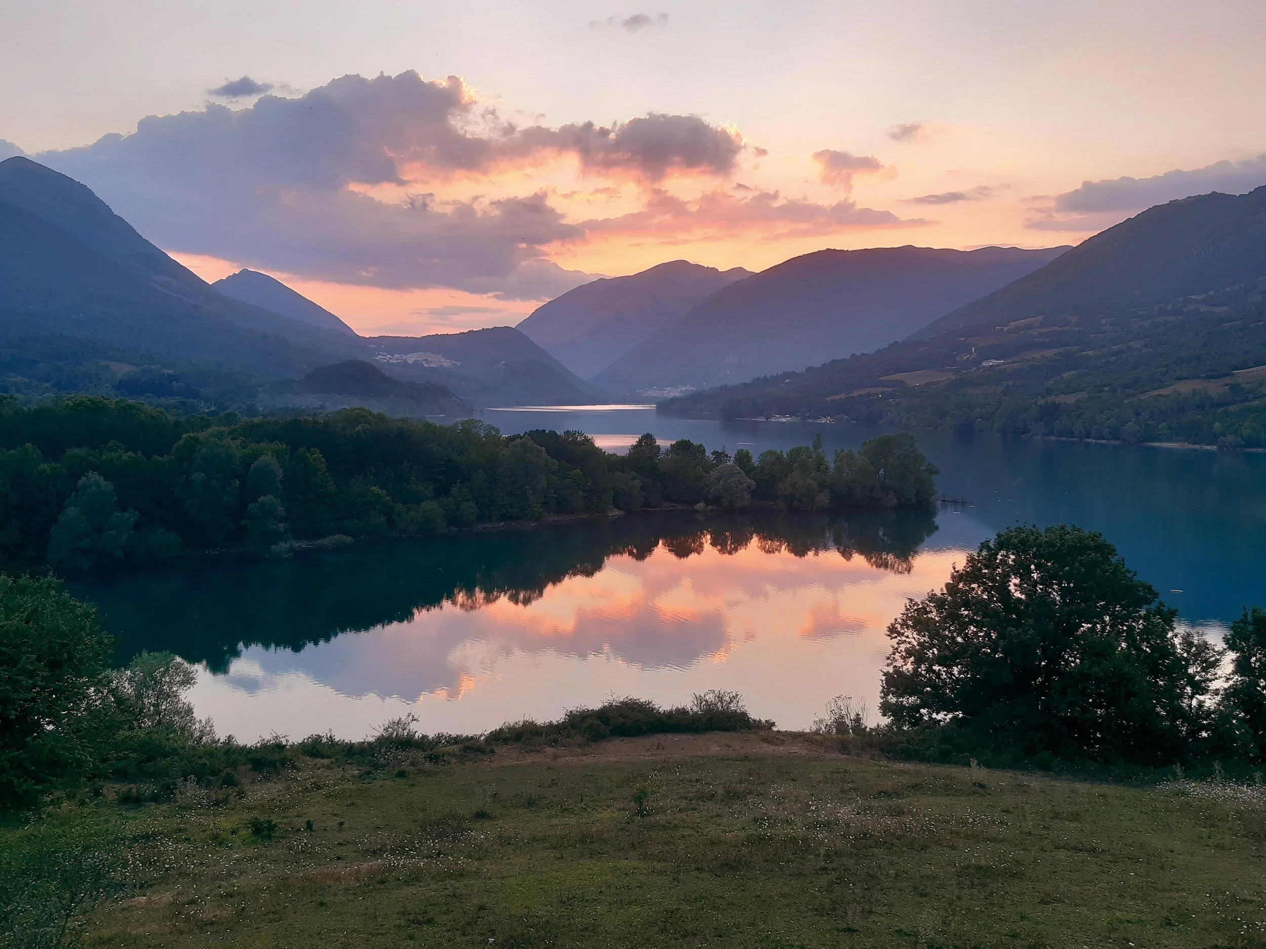 A tranquil lake surrounded by green trees and mountains at sunset, with colorful clouds reflected on the water.