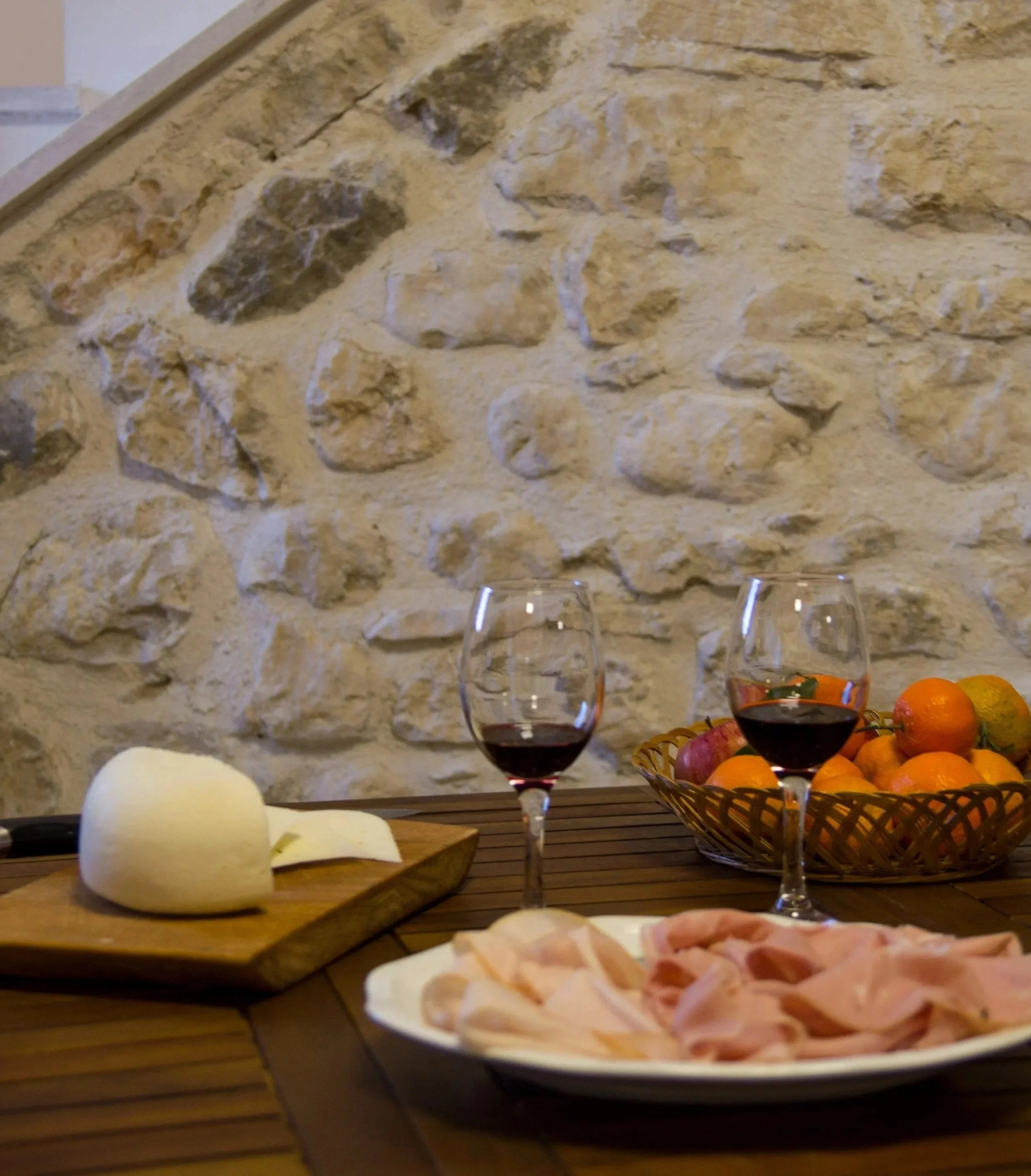 A table set with a plate of sliced ham, two glasses of red wine, a bowl of assorted oranges and apples, a block of cheese with a cheese knife, and a wooden serving board with a piece of cheese in front of a stone wall background.
