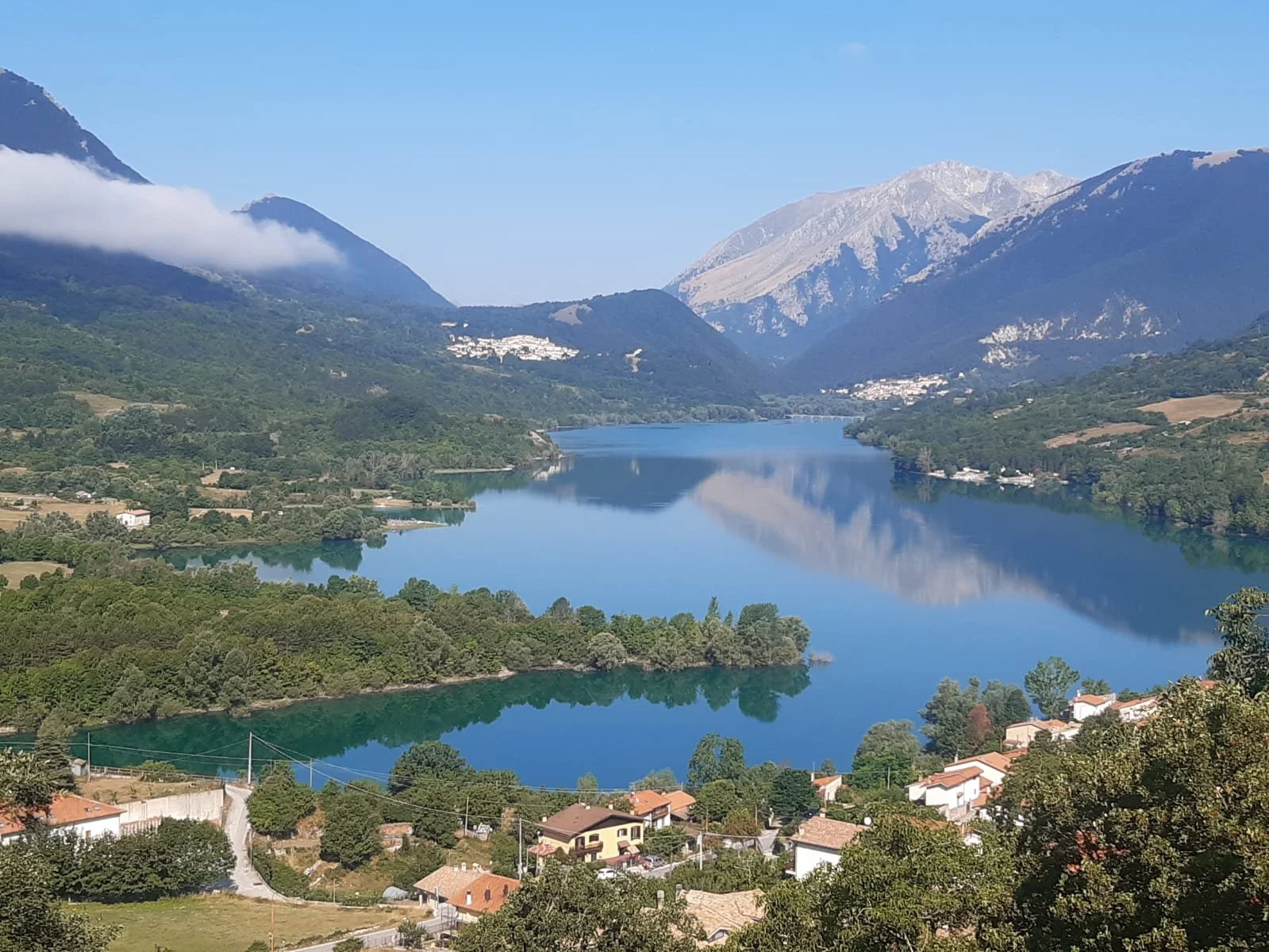 A scenic view of a winding river in a mountainous landscape with green hills, trees, and houses along the shore, under a clear blue sky.