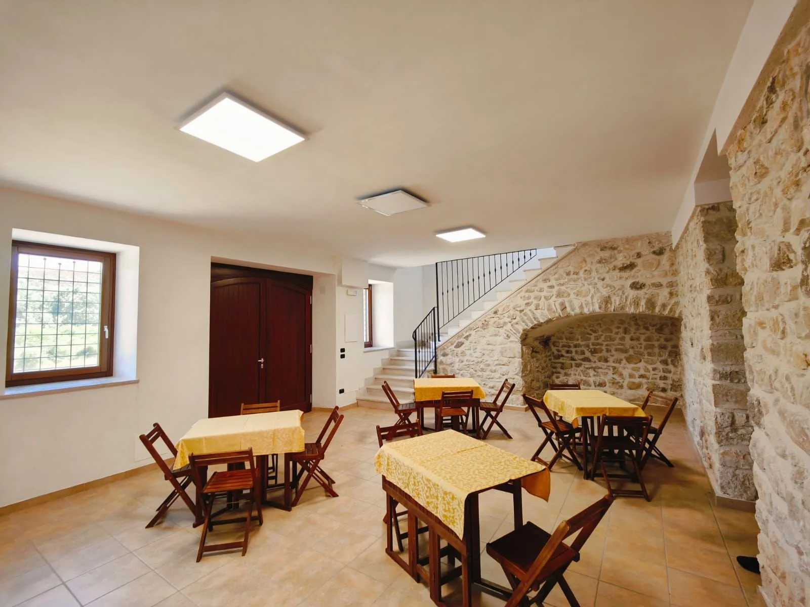Inside a cozy room with stone walls, wooden furniture, yellow tablecloths, metal staircase, and windows with natural light.