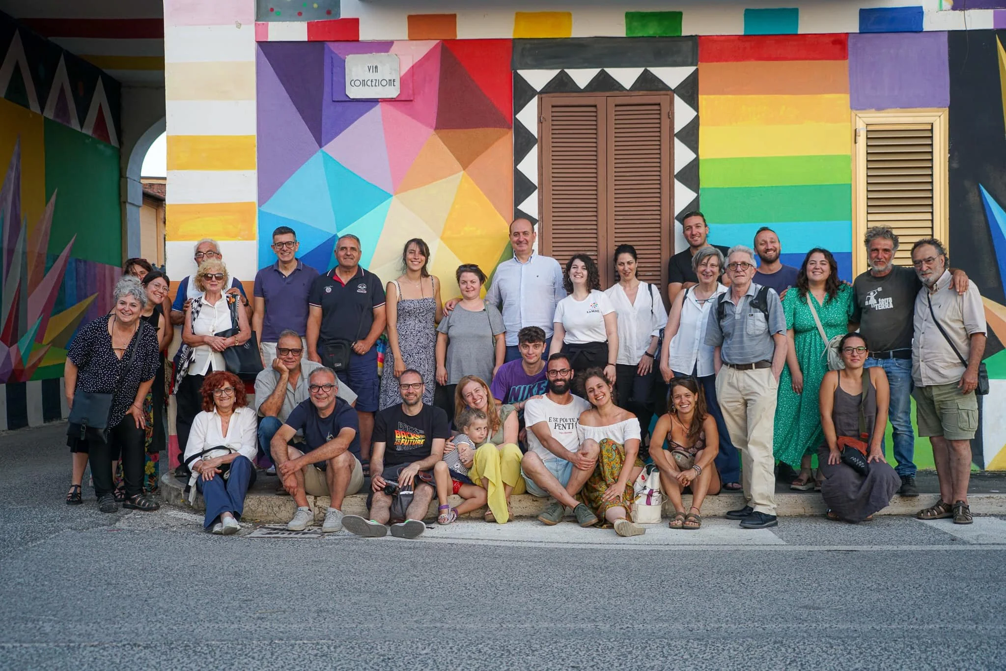 Group of people posing in front of a colorful geometric mural on a wall, some sitting on the curb and others standing behind them.