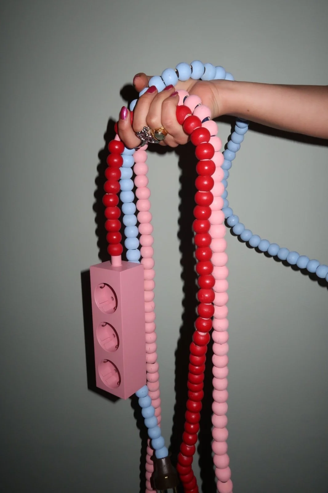 Person holding colorful beaded necklaces, including pink, red, and blue beads, with a pink power strip attached, against a plain wall.
