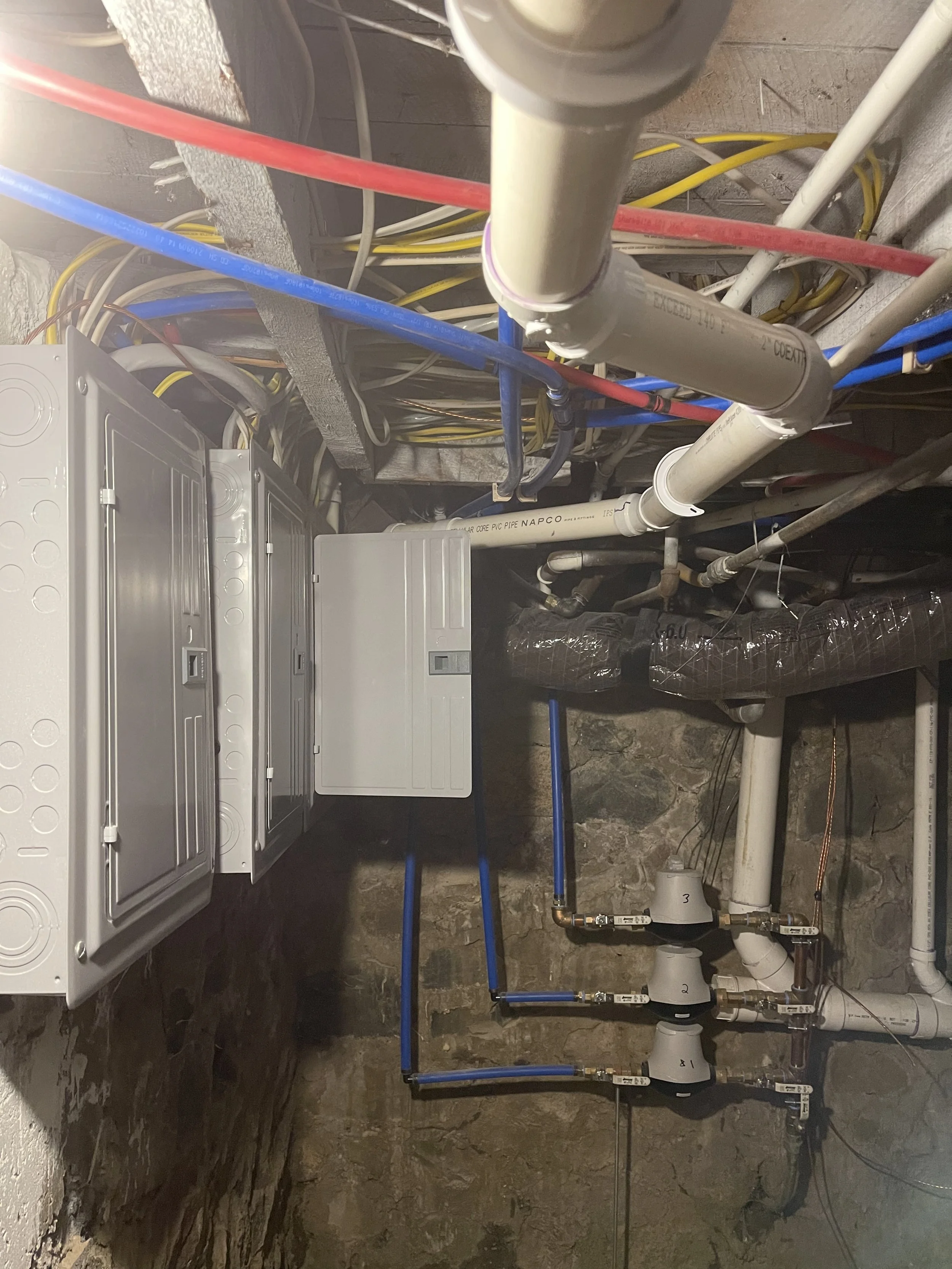 A basement utility room with various plumbing pipes, electrical panels, and wires, including colored pipes and control valves.