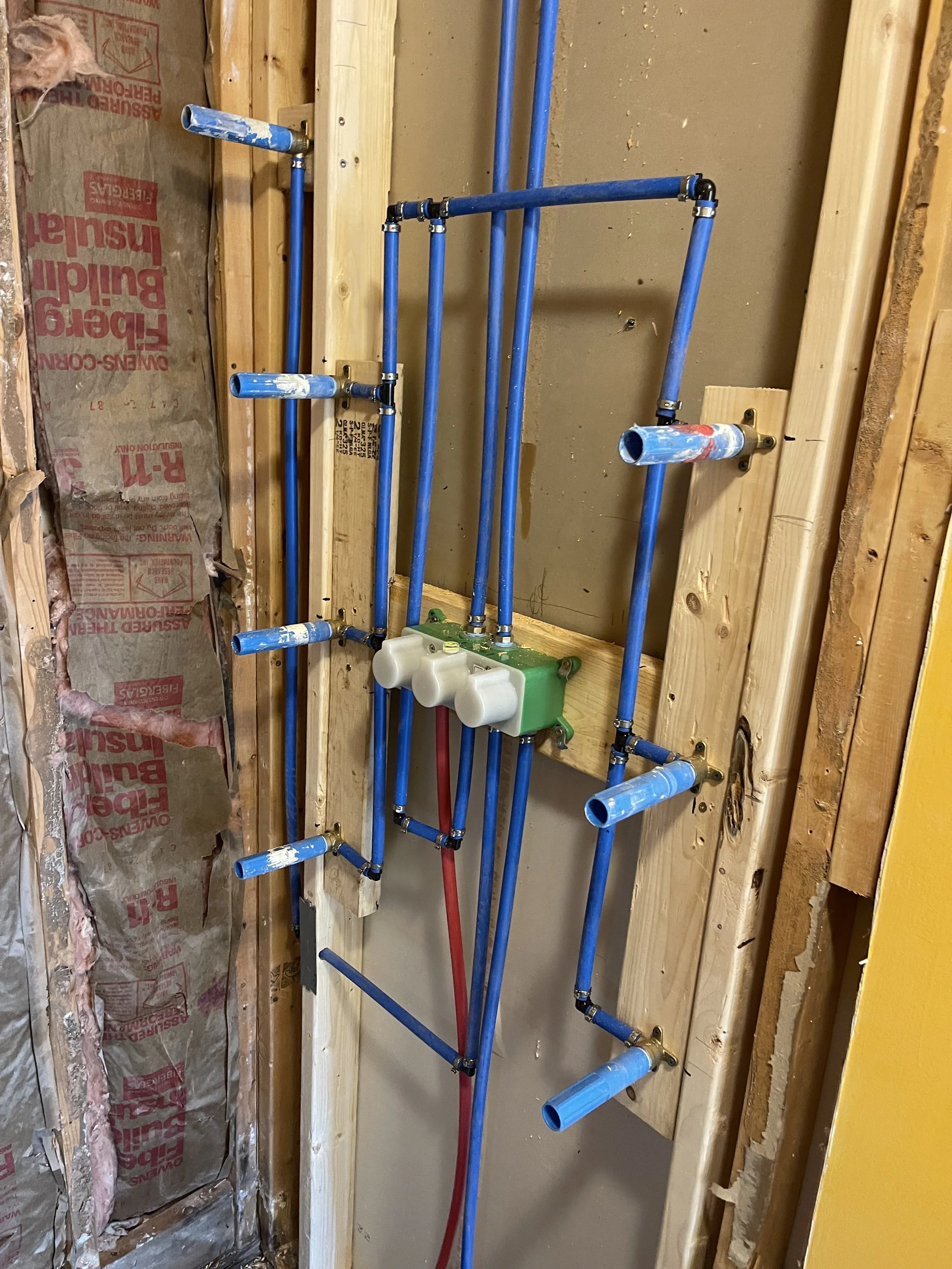 Blueprint of a plumbing wall setup with blue pipes, a green multi-valve component, and red and blue pipes connected to a wall framing.