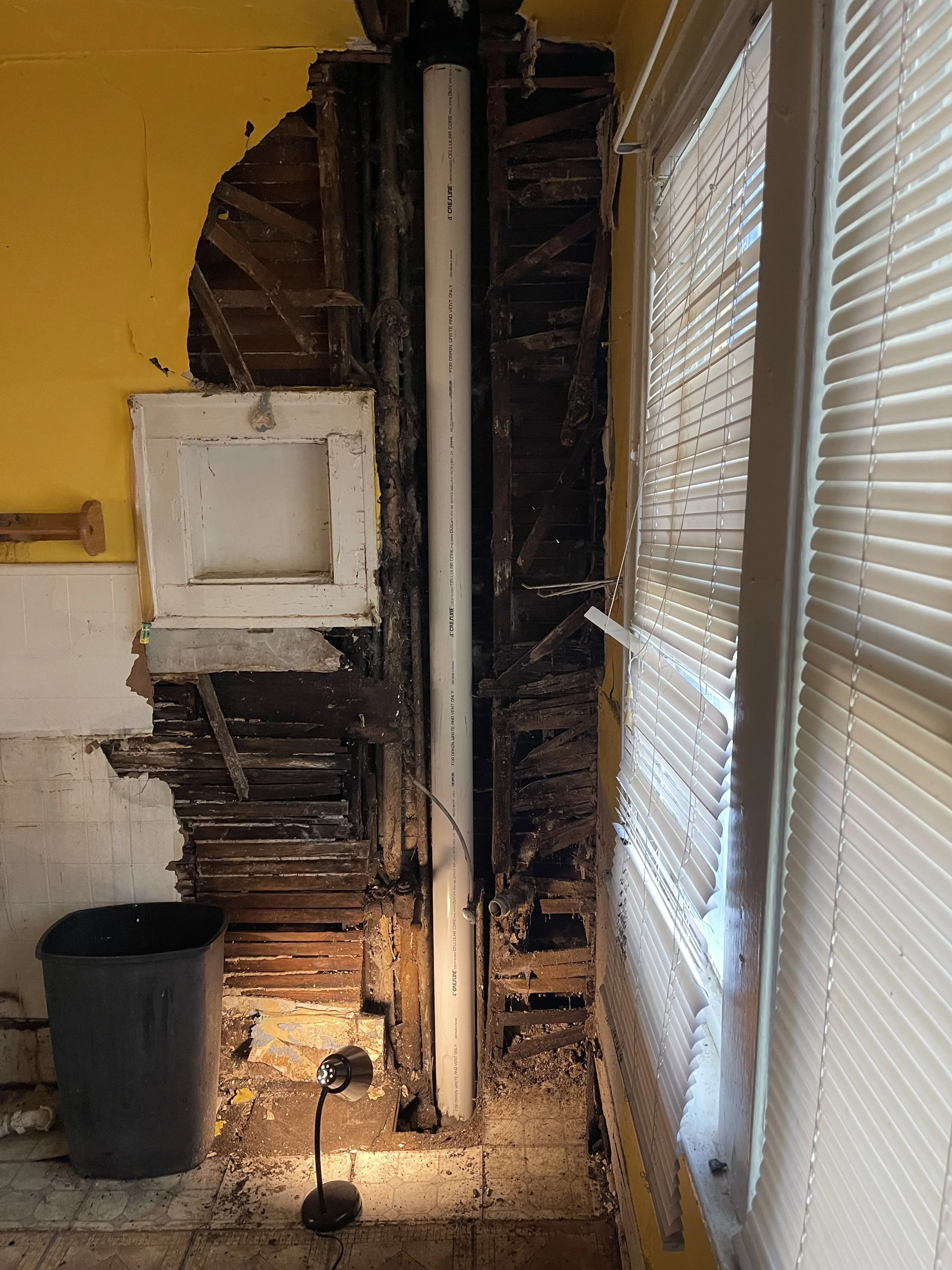 Room with exposed and damaged wall, partially removed drywall revealing wooden studs, a large pipe, a small white door frame, a black trash bin, a small desk lamp, and closed white window blinds.