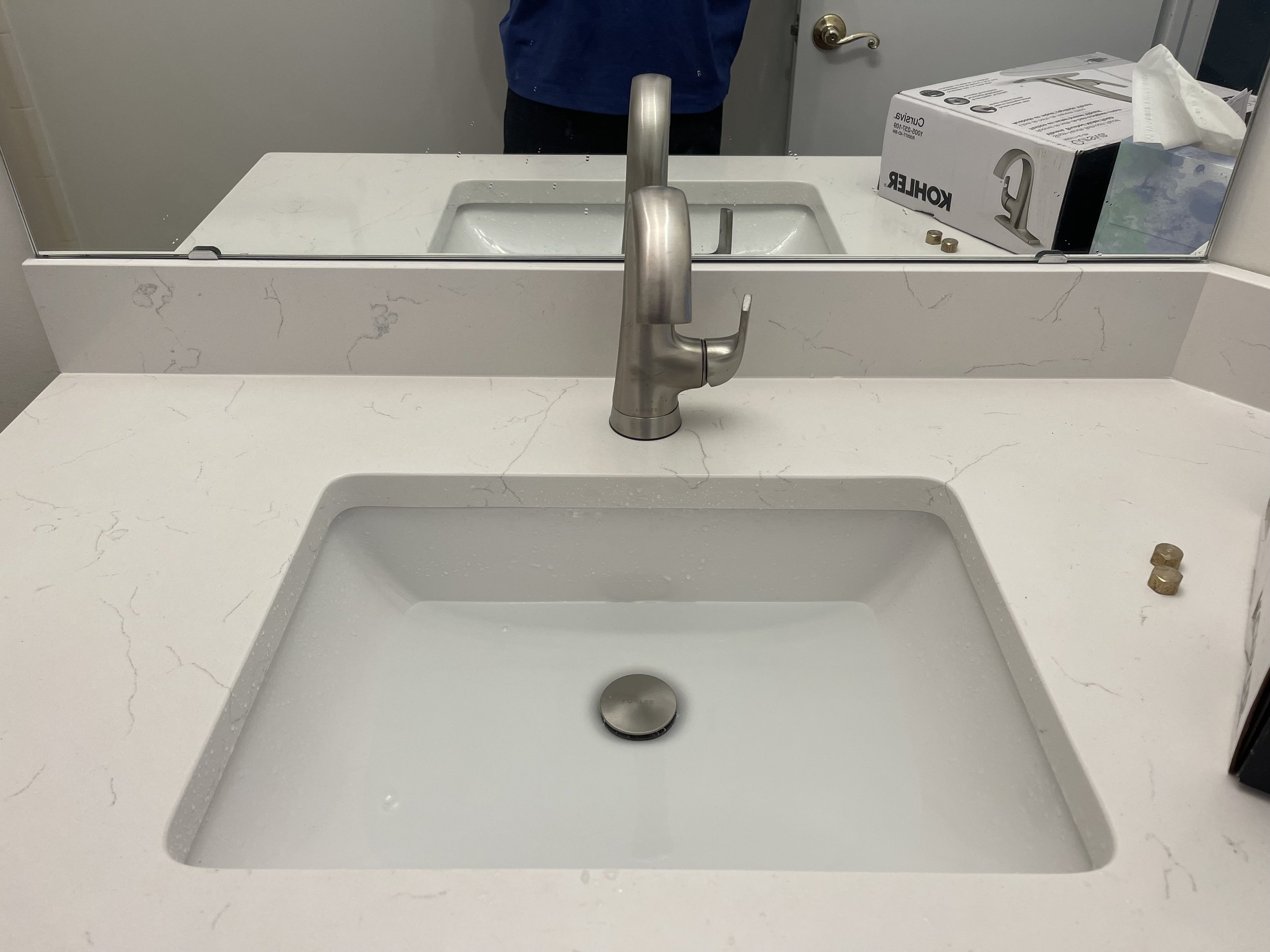 White marble bathroom sink with a silver faucet, a mirror, a box of Kohler parts, and two small brass fittings on the countertop.