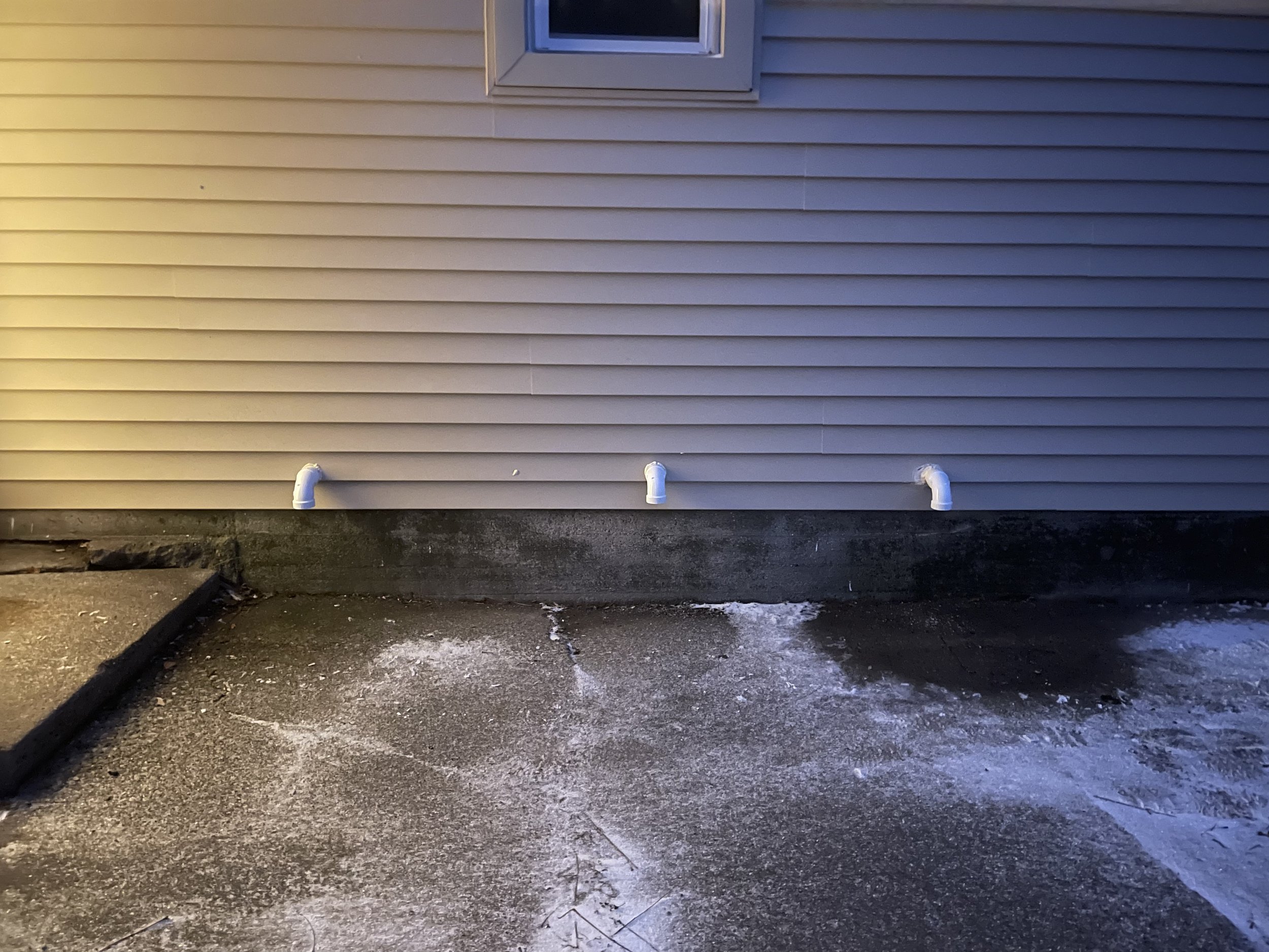 The exterior of a building with beige horizontal siding and three white pipes protruding from the bottom of the wall. The ground has patches of snow and ice.