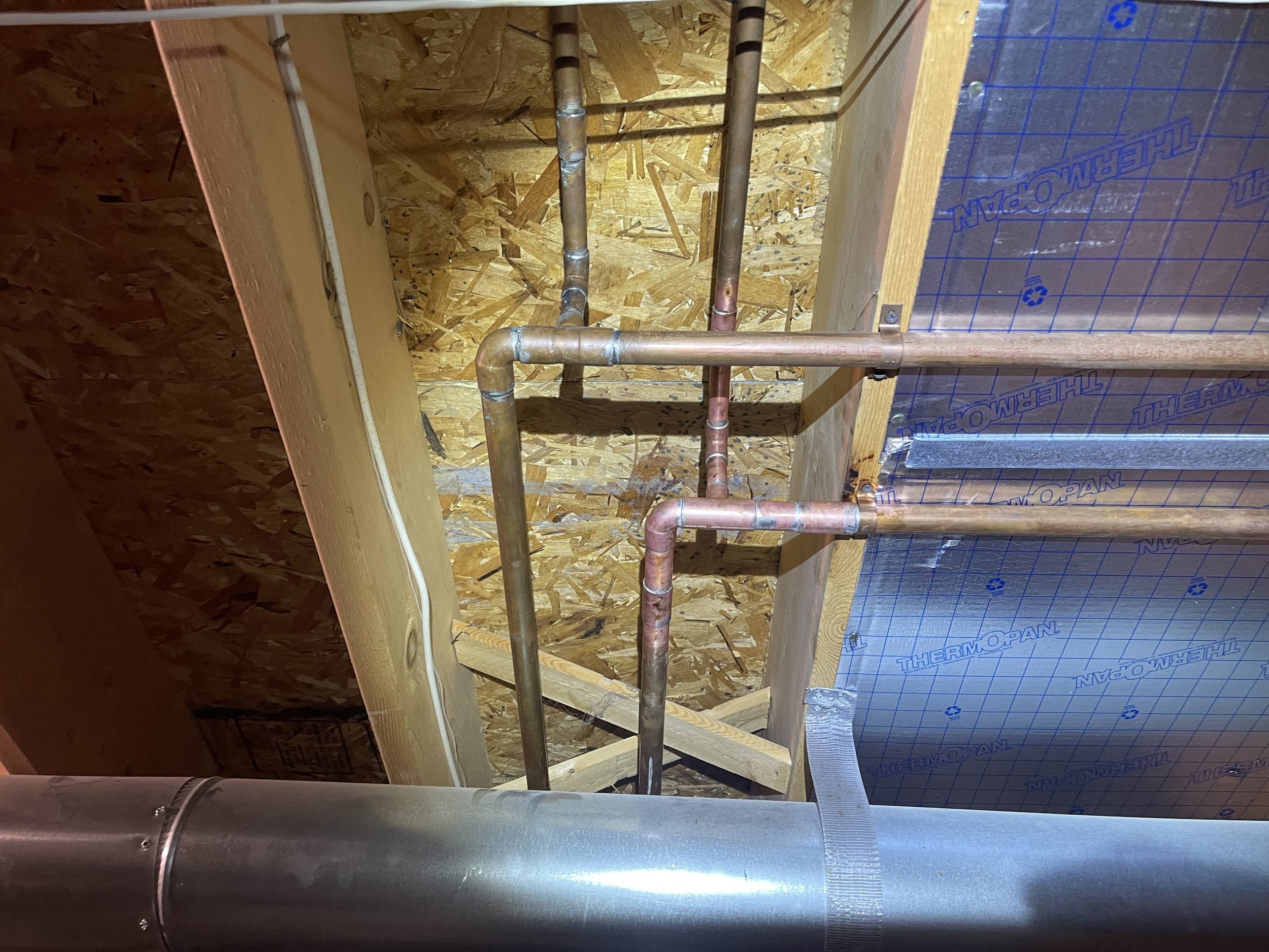 View of an unfinished ceiling with exposed plywood, insulation, and installed copper and metal pipes.