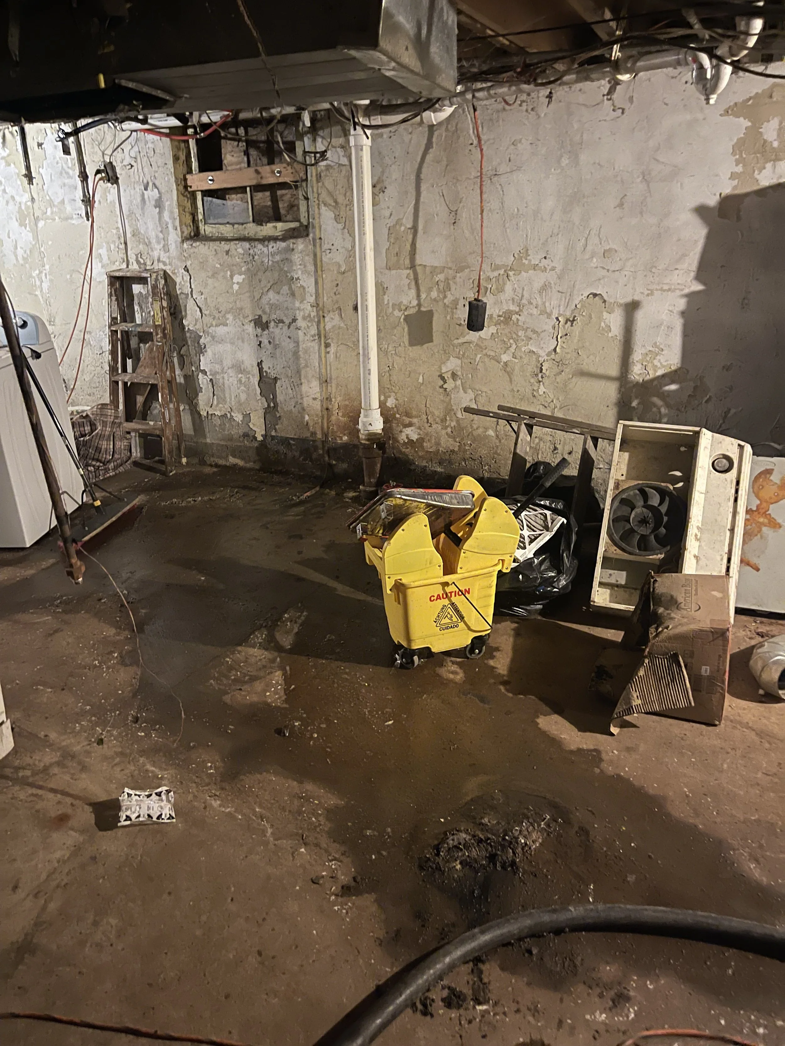 Construction or renovation site with a wet concrete floor, a yellow caution mop bucket, a small ladder, a fan attached to a vent, and an old washing machine, with exposed pipes and peeling paint on the walls.