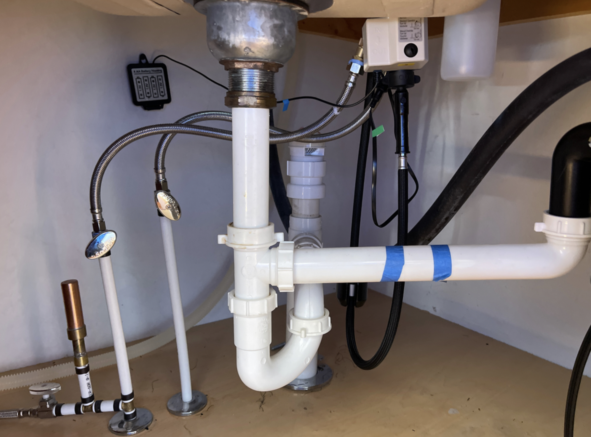 Underneath a sink showing plumbing pipes, hoses, and electronic components connected to the plumbing system.