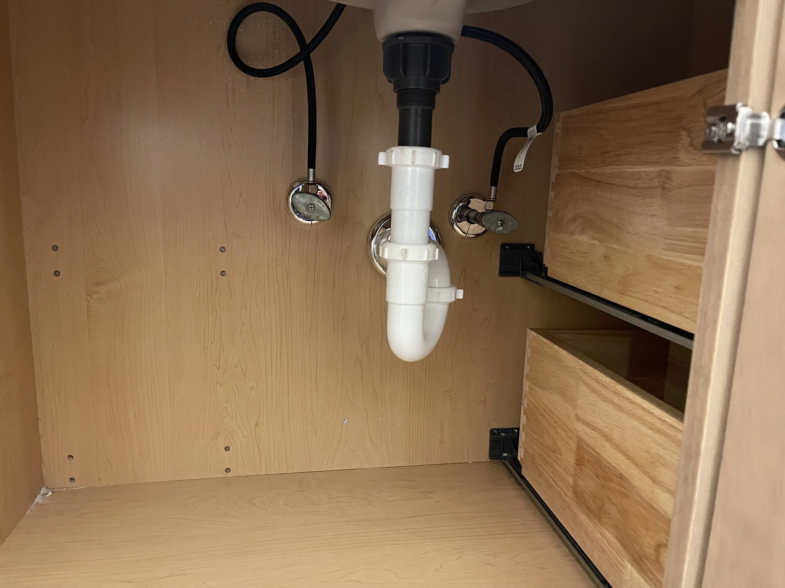 View of the inside of a wooden kitchen cabinet, showing plumbing pipes under the sink and two wooden drawers with black handles on the right side.