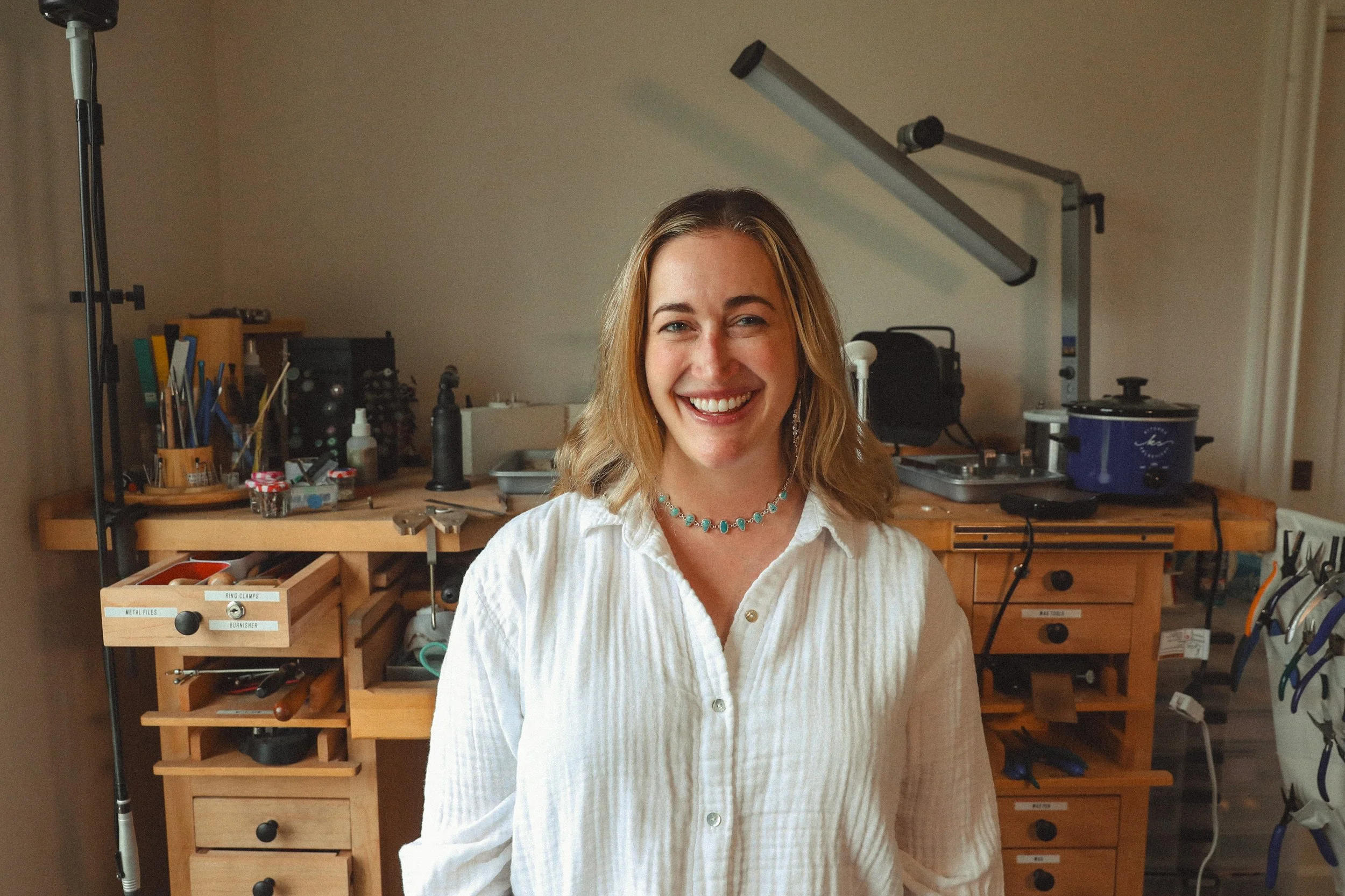 Beach scene with white umbrellas, beach chairs with white cushions, and a small wicker table on sand near the ocean, rocks, and waves.