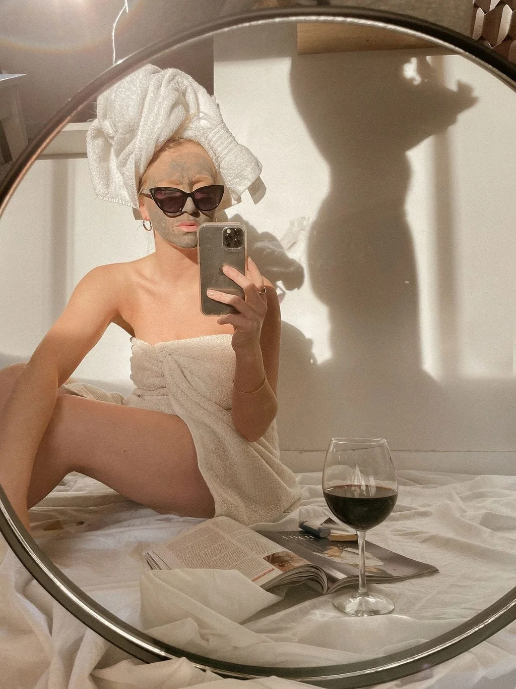 Person with a towel on their head, sunglasses, and a face mask taking a mirror selfie in a bedroom, with a glass of red wine and an open magazine on the bed.