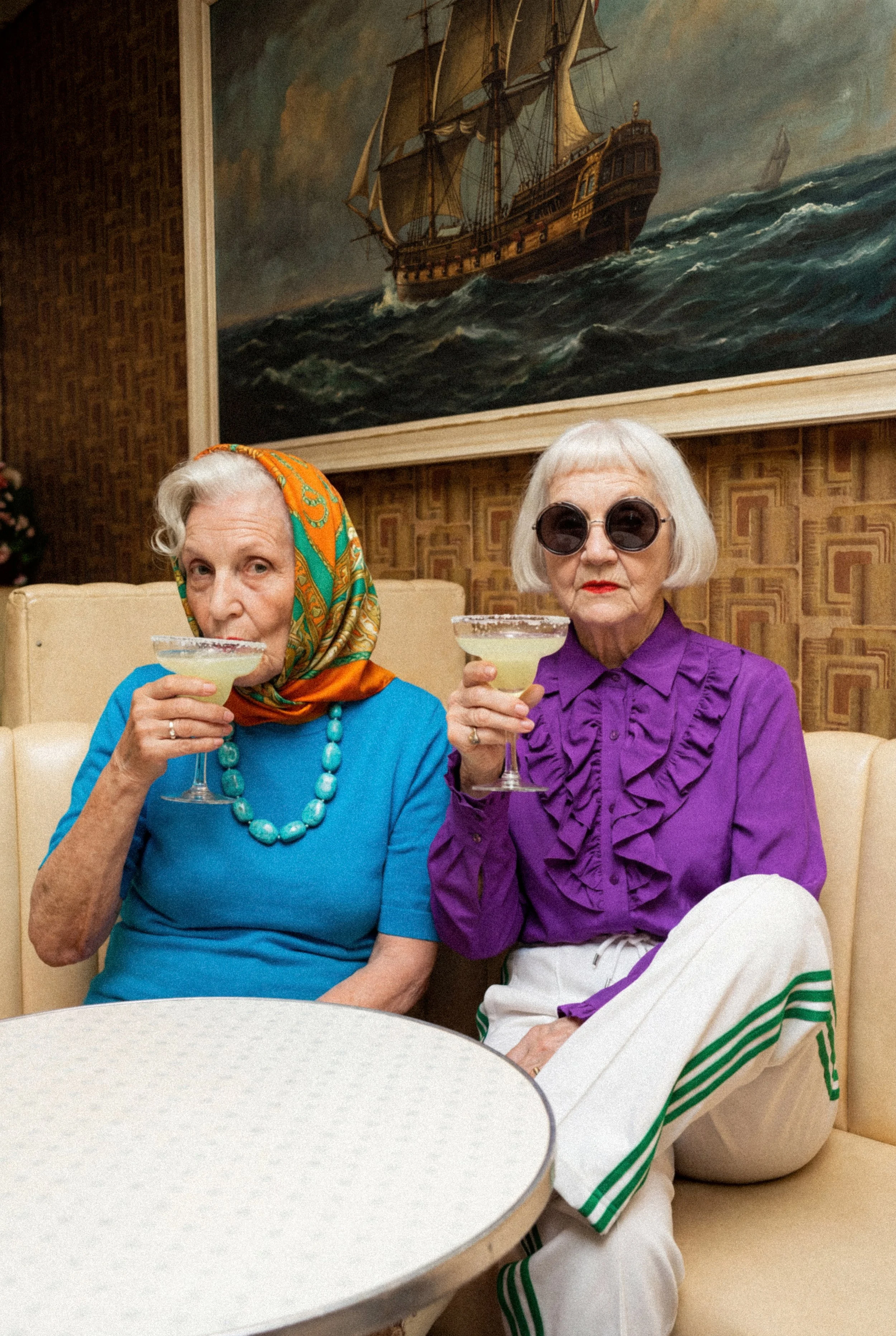 Two elderly women sitting on a beige couch, holding margarita glasses and sipping drinks, with a large maritime painting of a sailing ship hanging on the wall behind them.
