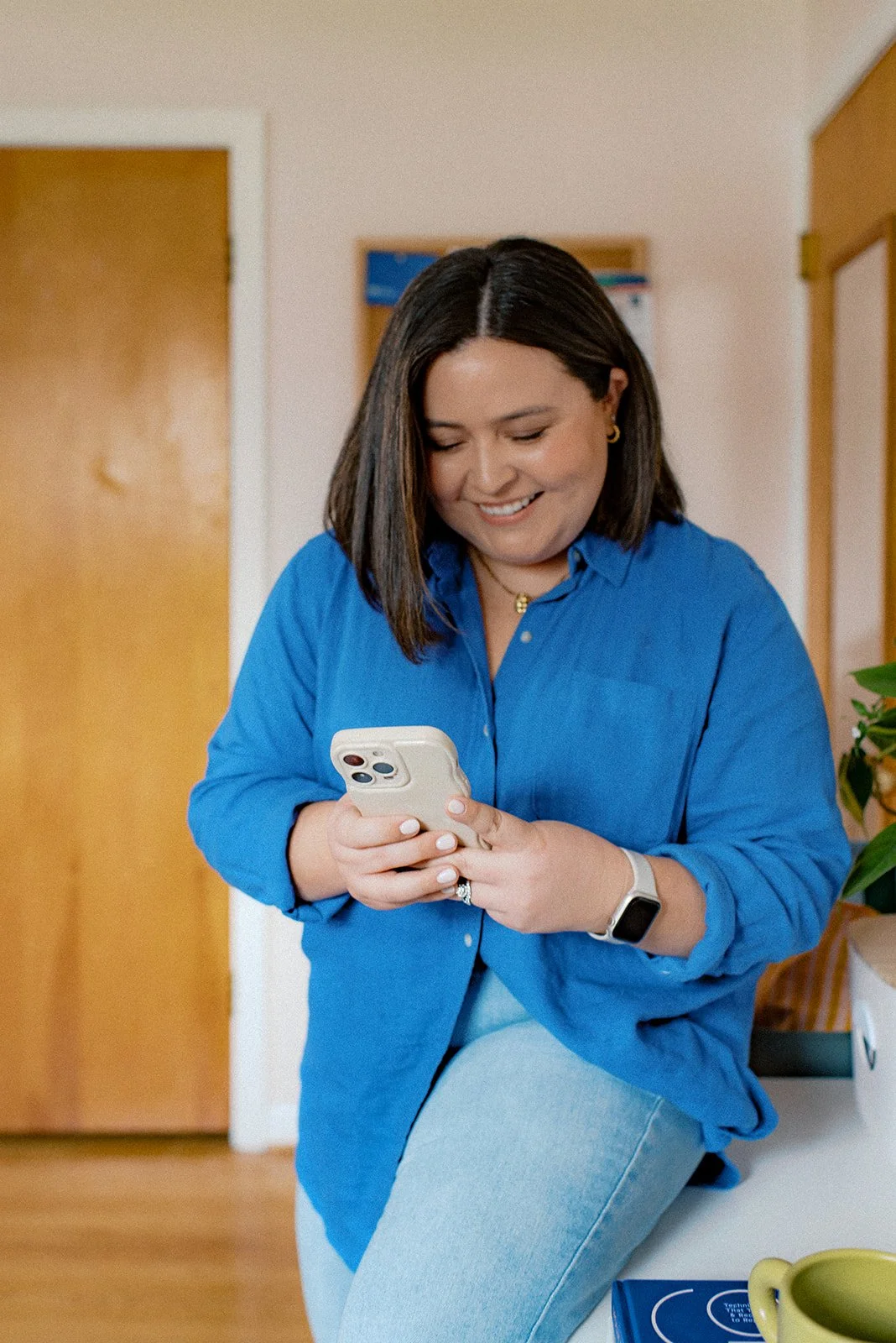 A woman with shoulder-length dark hair, wearing a blue button-down shirt and light jeans, smiling while looking at her smartphone. She has a smartwatch on her left wrist and gold jewelry, and is sitting on a white surface in a room with wooden doors and a potted plant nearby.