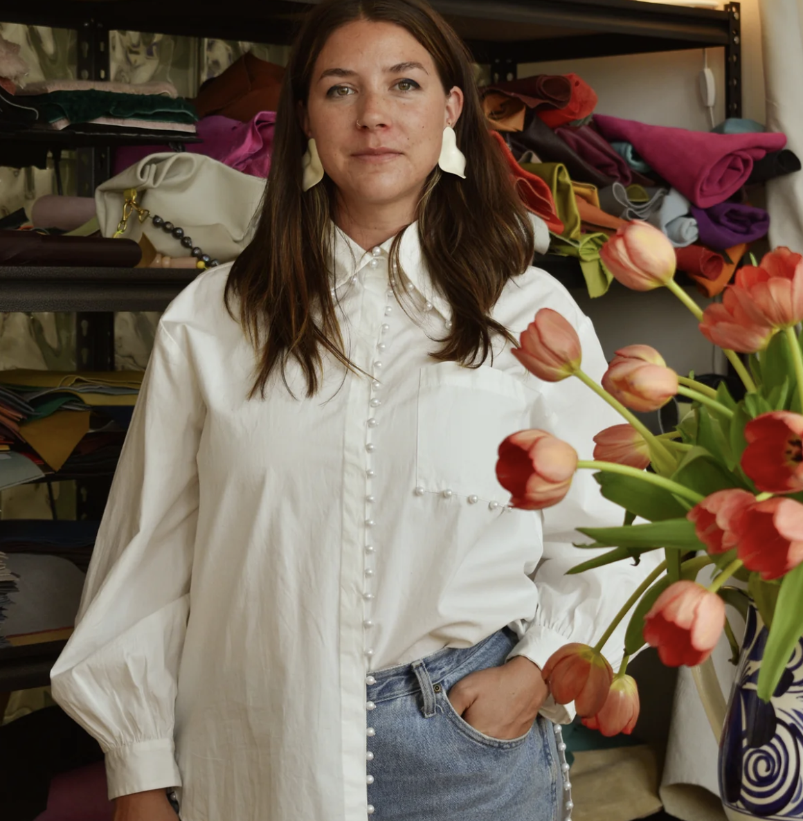 A young woman with dark hair smiling and looking upward, showing her teeth, wearing gold earrings and a chain necklace, with her hand resting on her face.