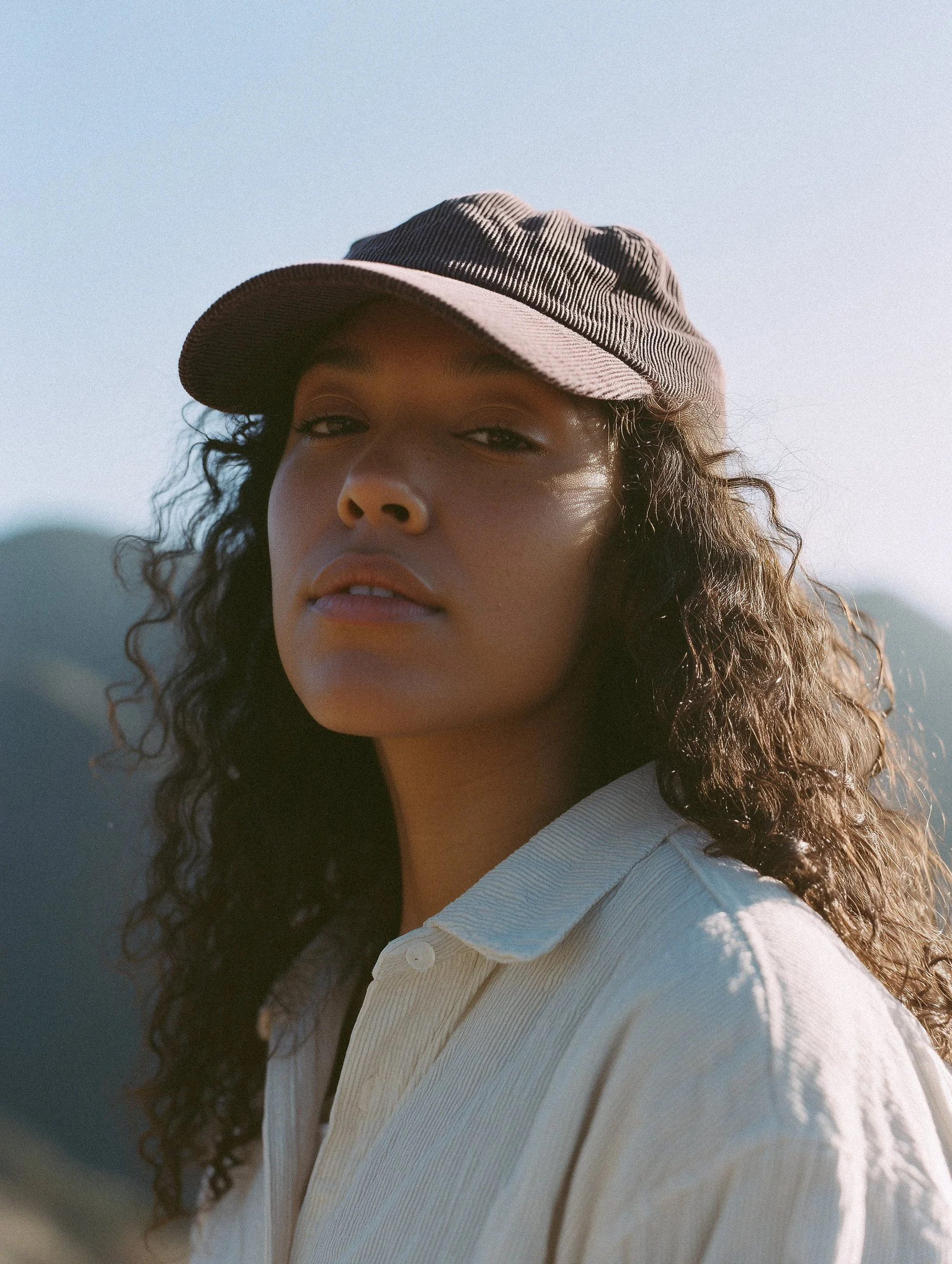 A young woman with curly hair wearing a beige cap and a light-colored shirt outdoors near mountains.