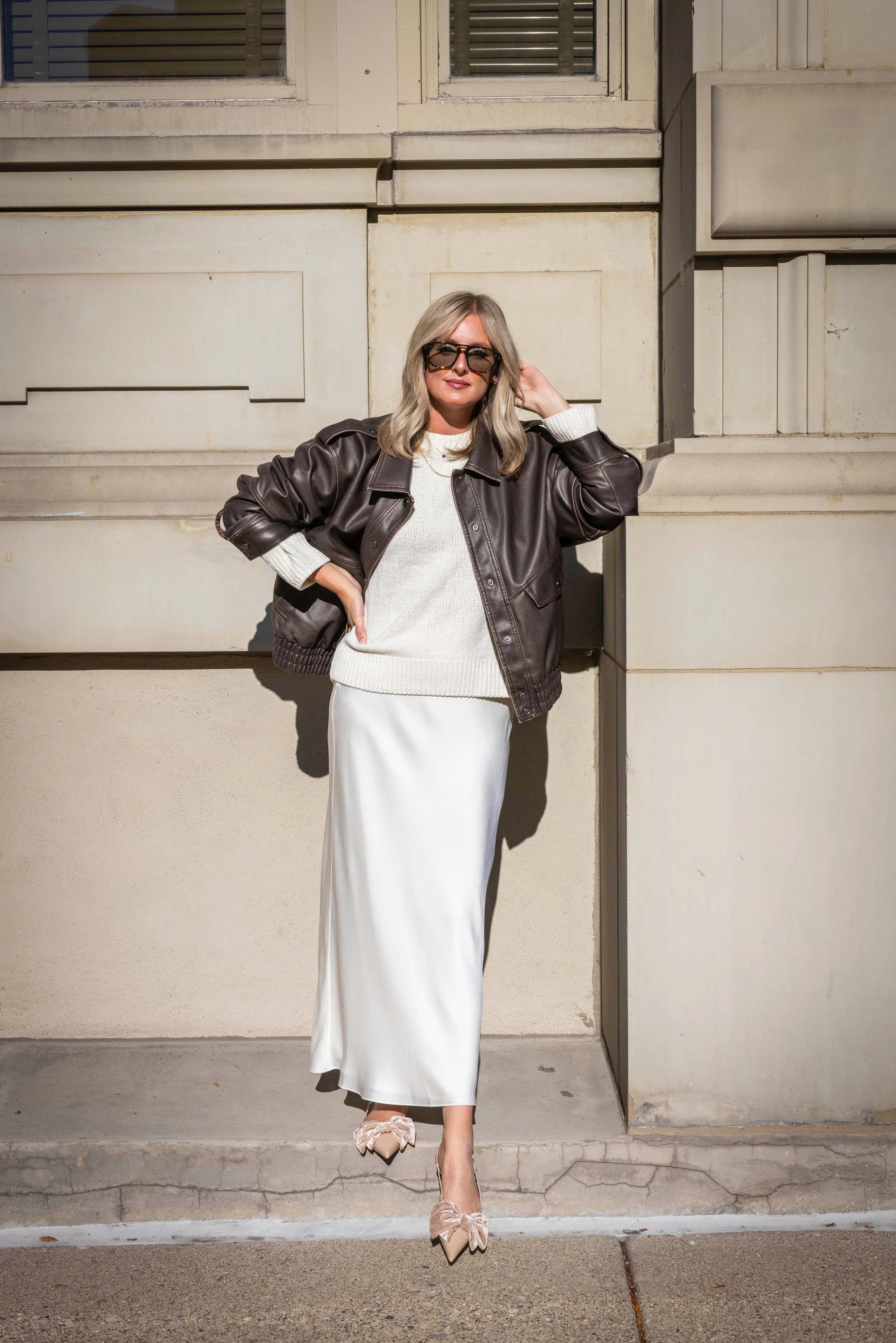 A woman with blonde hair and sunglasses standing outside a light-colored building wall, wearing a white sweater, white skirt, brown leather jacket, and beige high heels with bows.