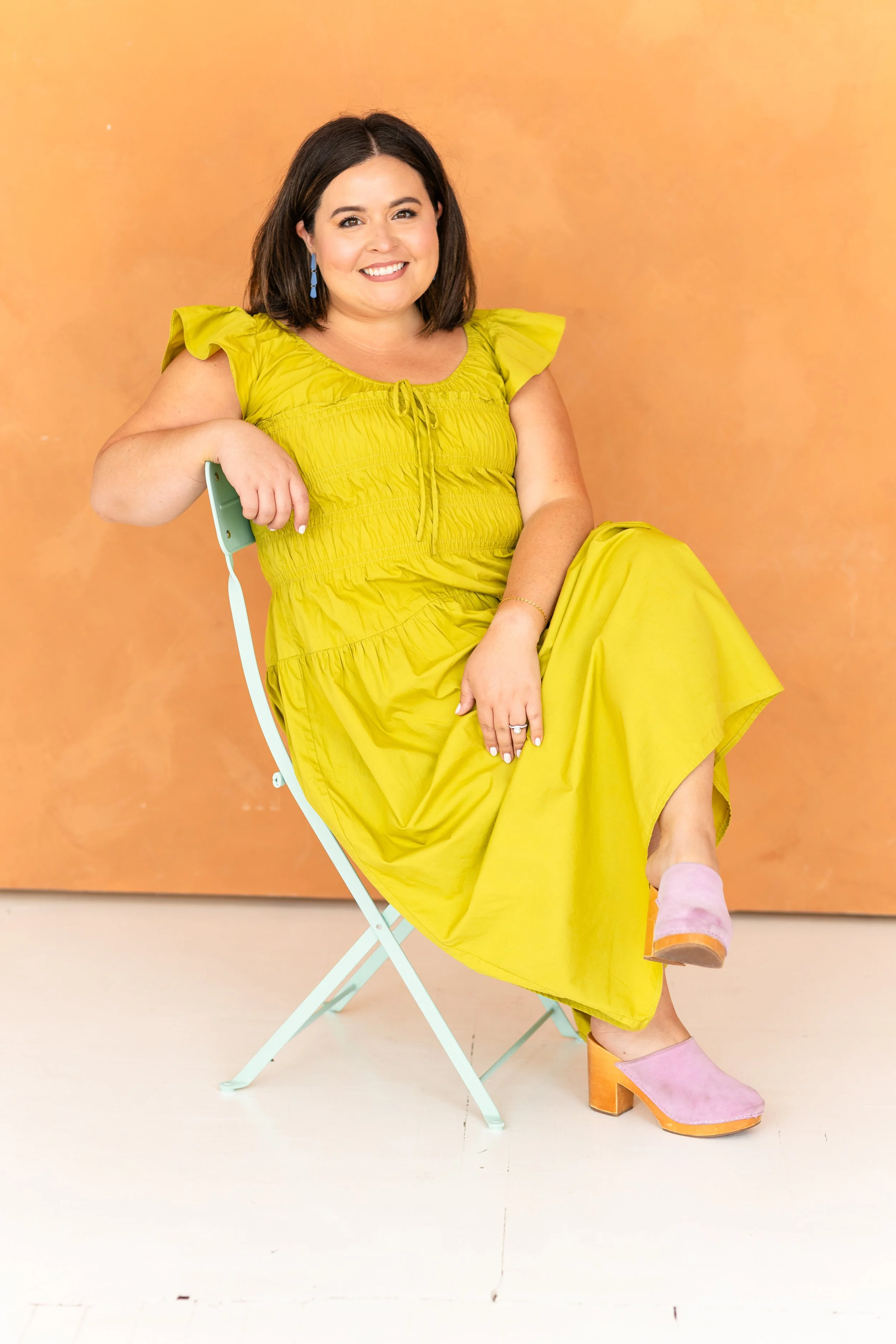 Woman sitting on a pastel chair against an orange background, wearing a yellow dress and pink shoes, smiling at the camera.