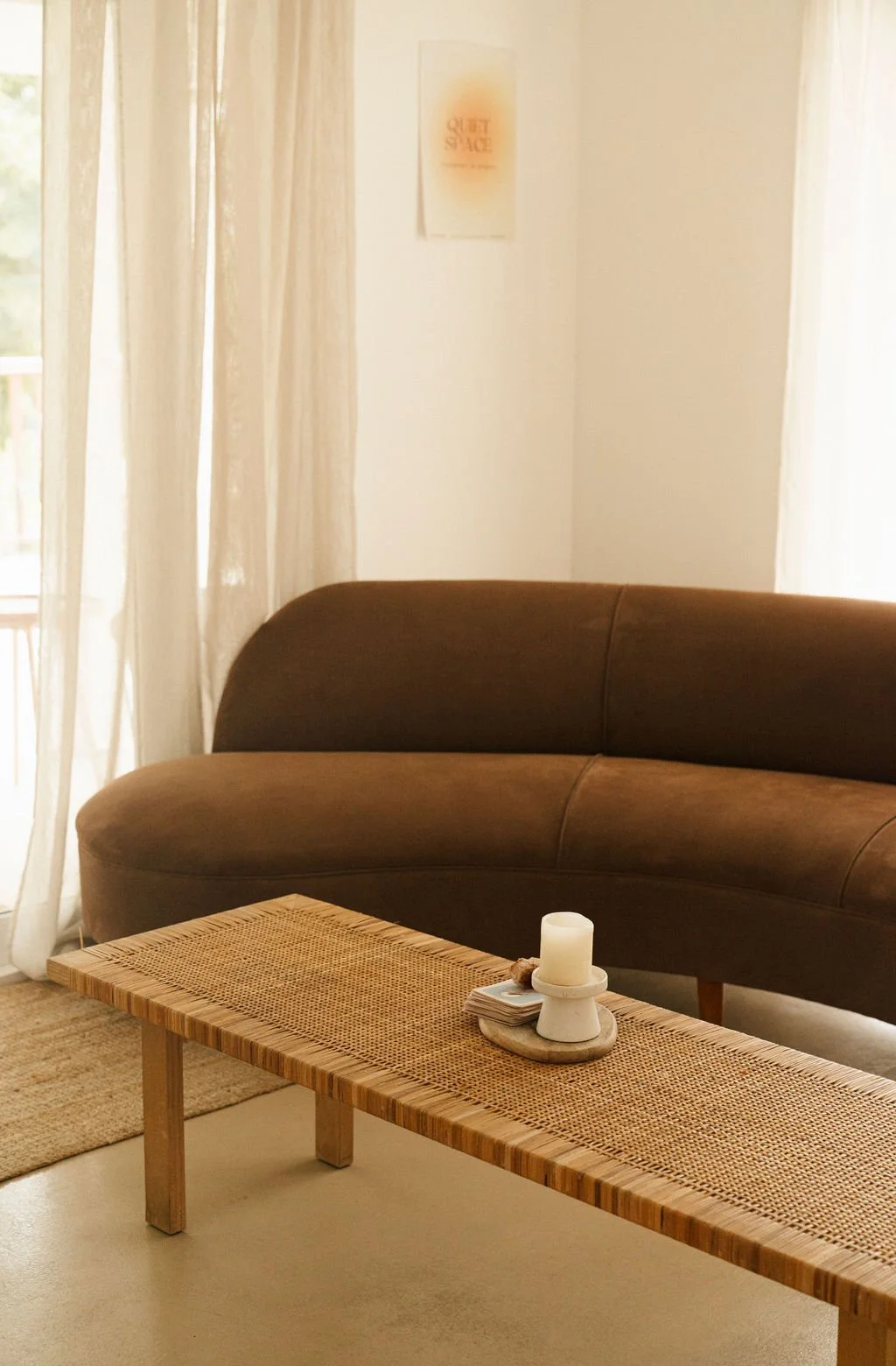 A cozy living room with a rattan coffee table, a small candle and a deck of cards on top, a brown velvet sofa, and cream curtains letting in natural light.