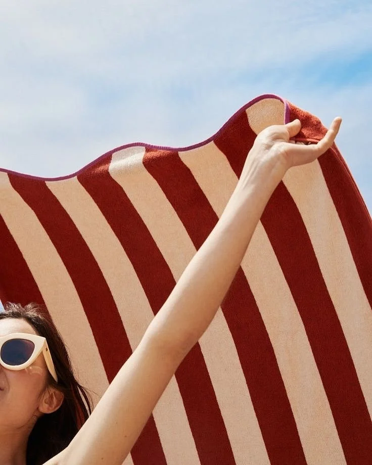 A woman wearing sunglasses lying under a large red and white striped beach umbrella, looking up at the sky.