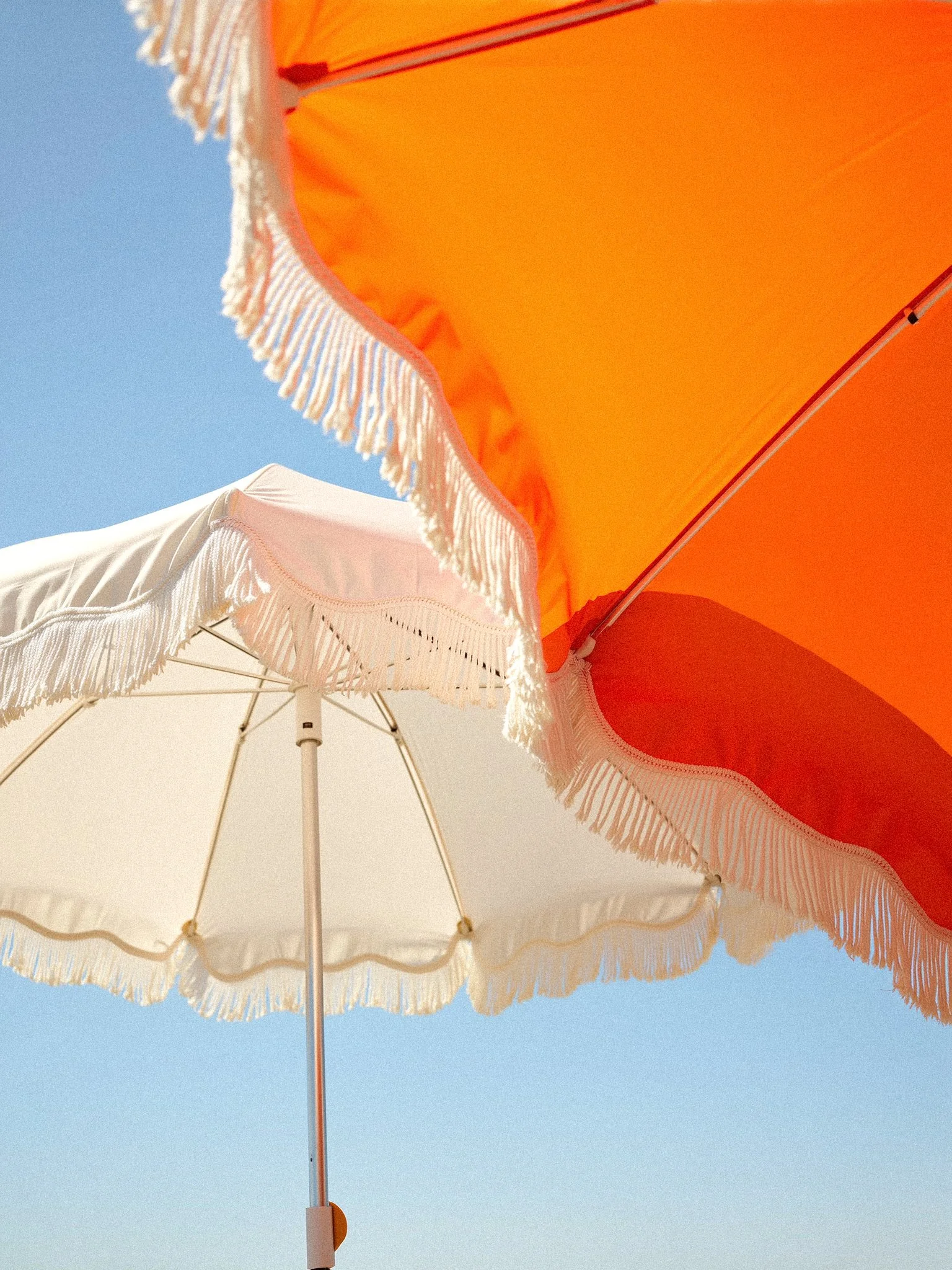 View of two beach umbrellas, one white with fringe and the other orange with fringe, against a clear blue sky.