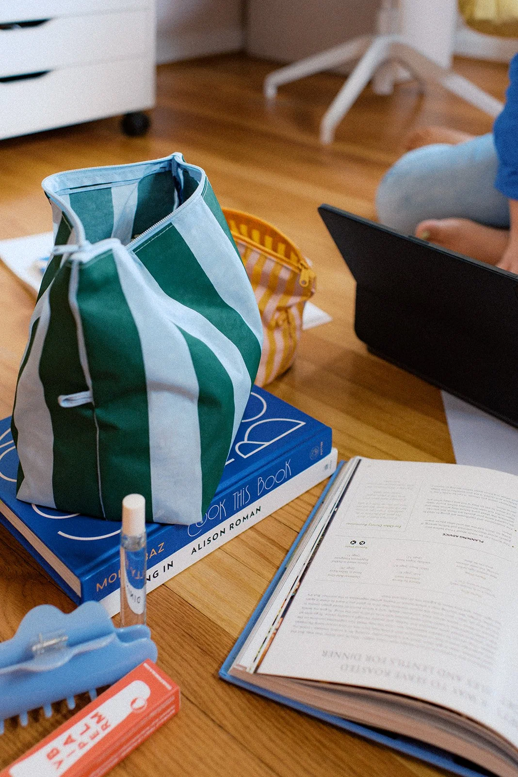 A desk with a large blue and green striped tote bag placed on top of a blue book titled 'Look This Book,' a pen, an open notebook or magazine, and a black laptop. In the background, part of a person sitting on the floor is visible.