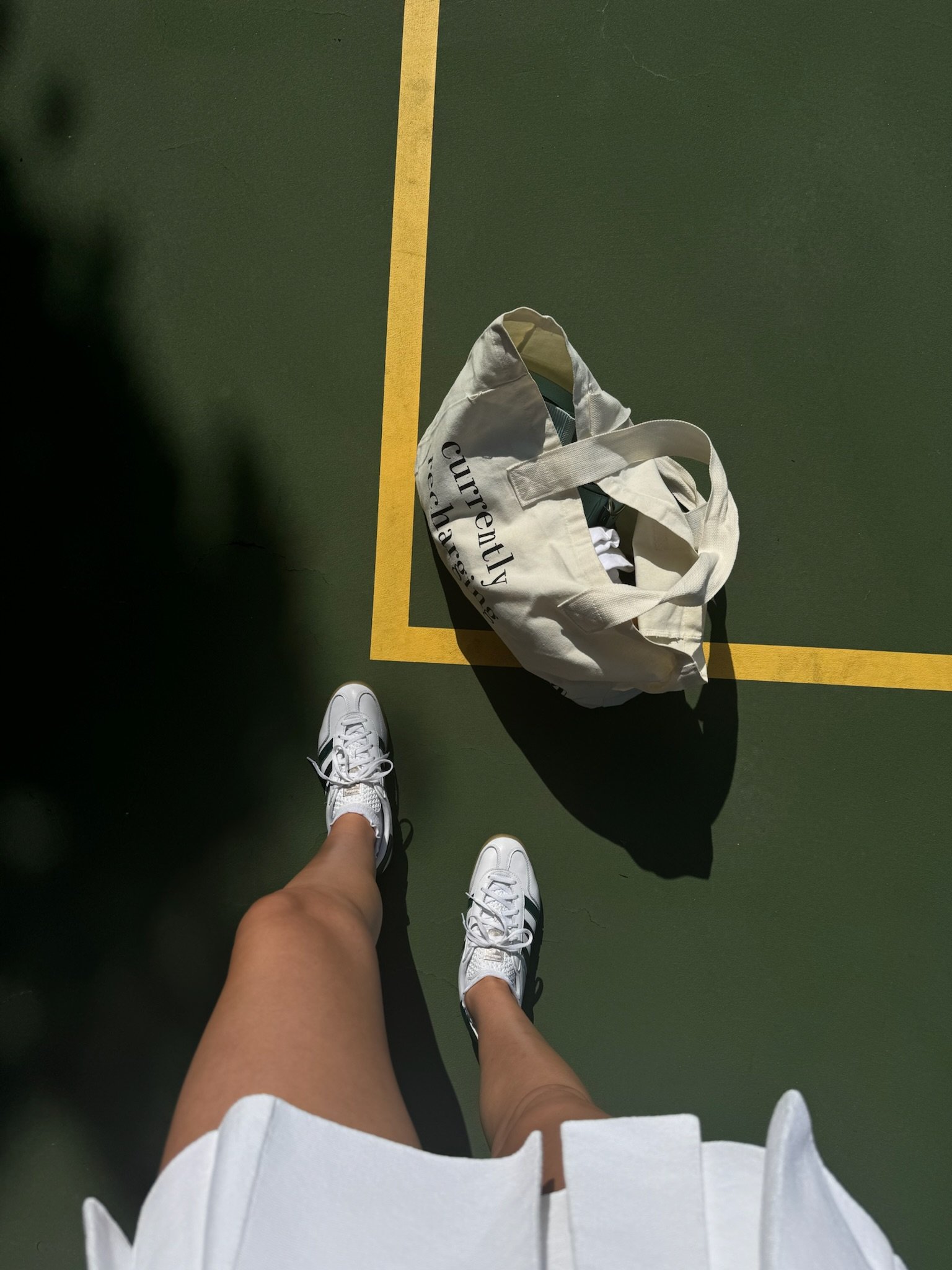 Top-down view of a person standing on a tennis court, wearing white sneakers and a white skirt, with a tote bag on the ground nearby.