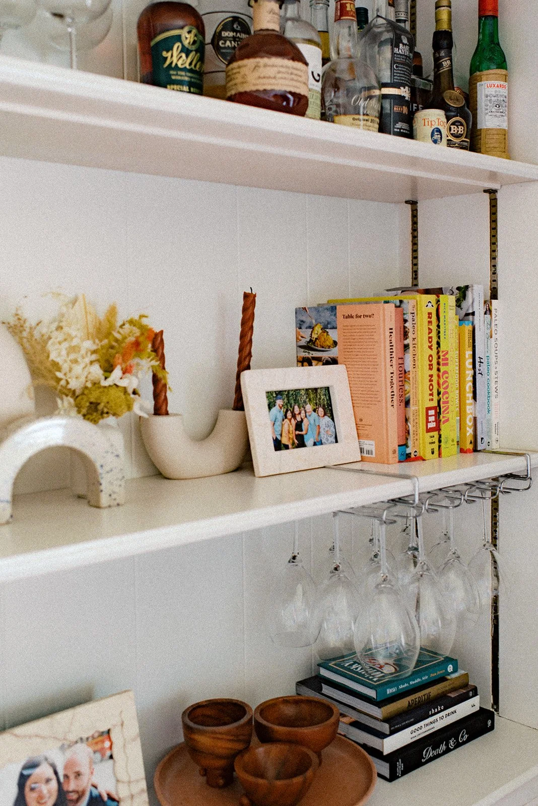 White shelves holding liquor bottles, cookbooks, a framed photo, glassware hanging upside-down, wooden bowls, and a small framed picture.