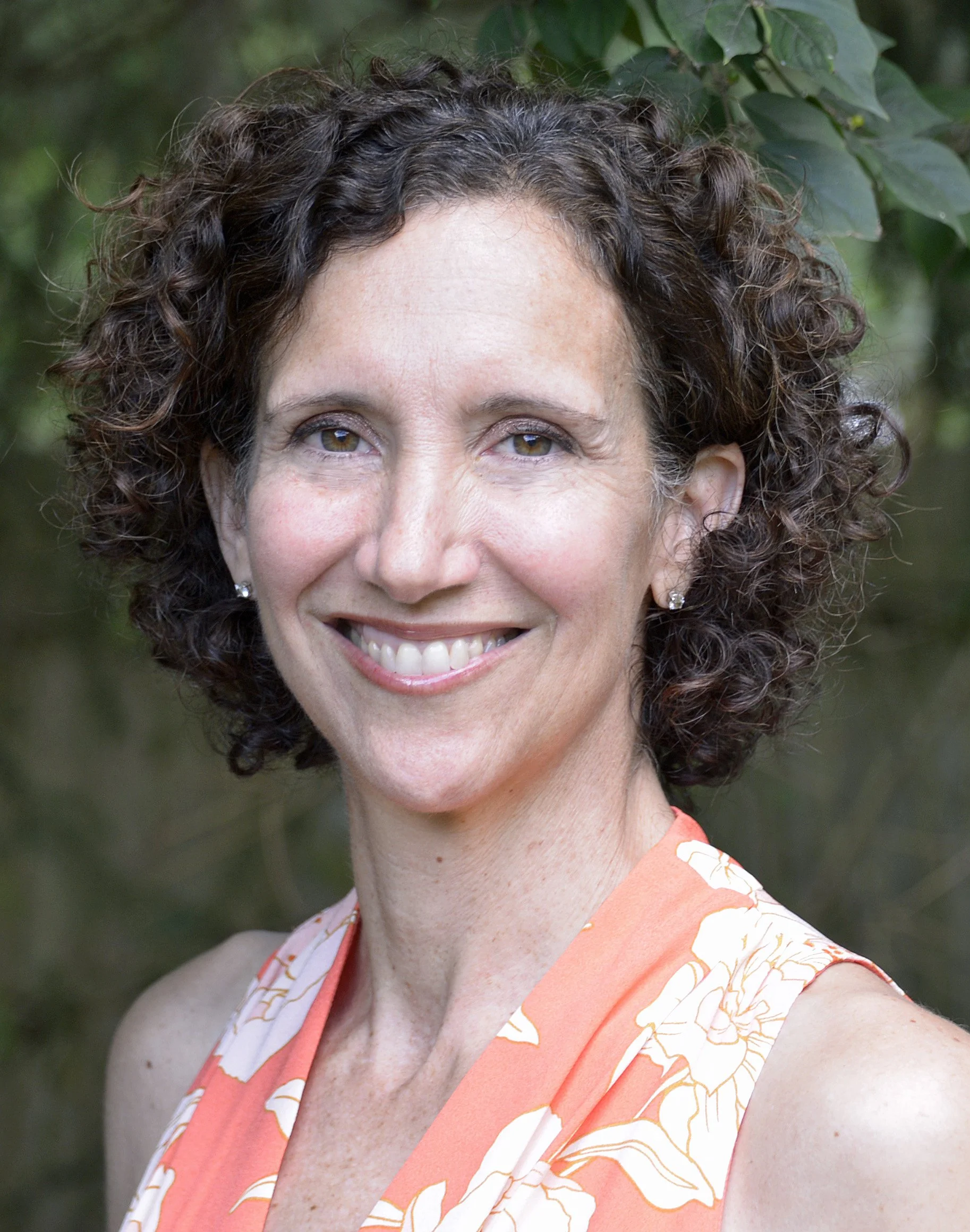 A woman with curly brown hair smiling outdoors, wearing a sleeveless orange dress with a floral pattern and earrings.