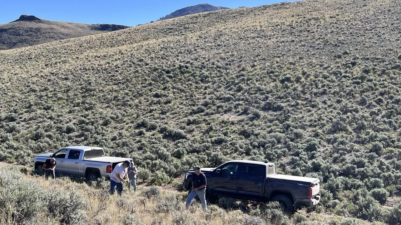 Four people standing near two pickup trucks on a hillside covered with shrubs and dry grass, with mountains in the background under a clear blue sky.